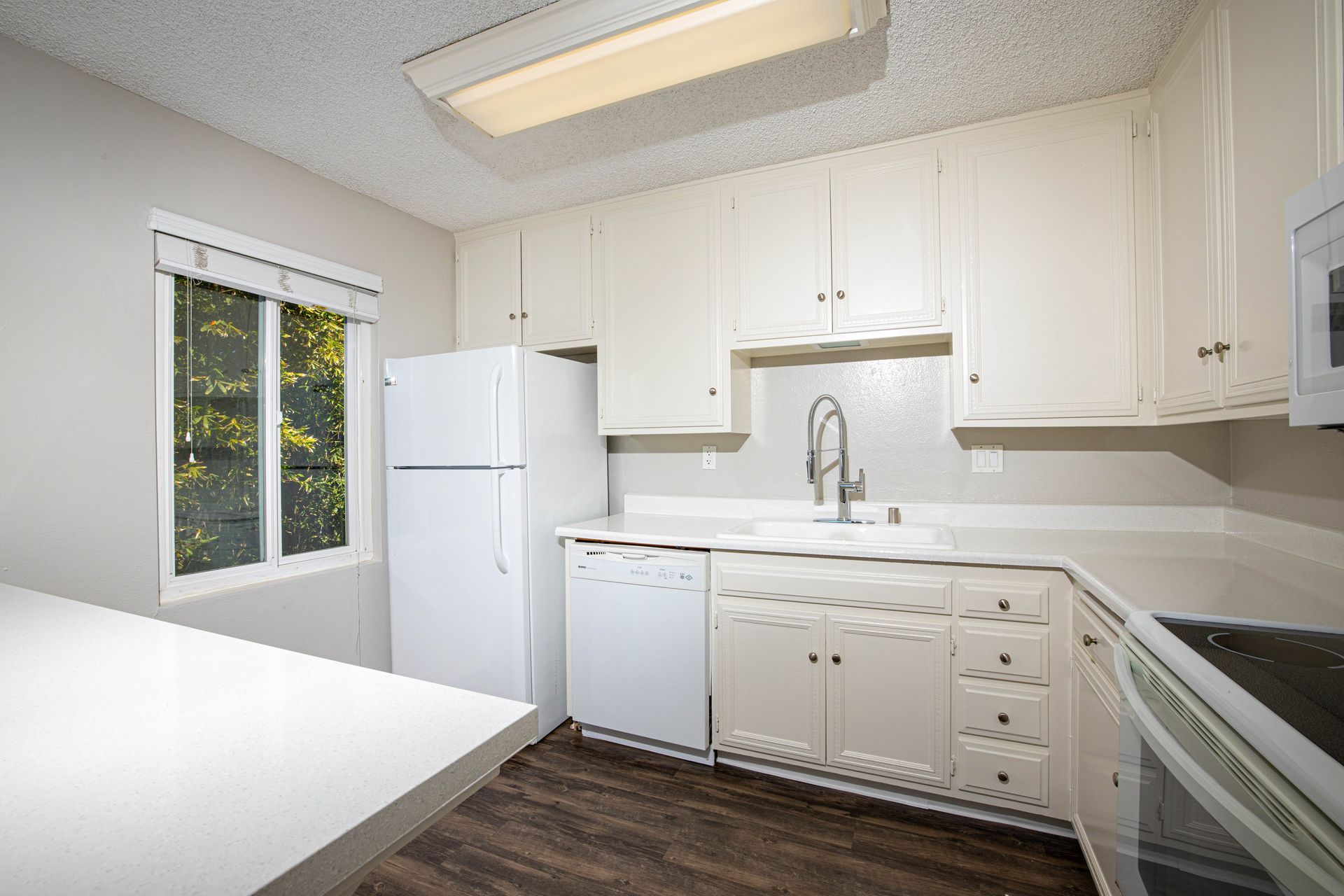 Bright white kitchen with cabinets, refrigerator, sink, stove, and window overlooking greenery