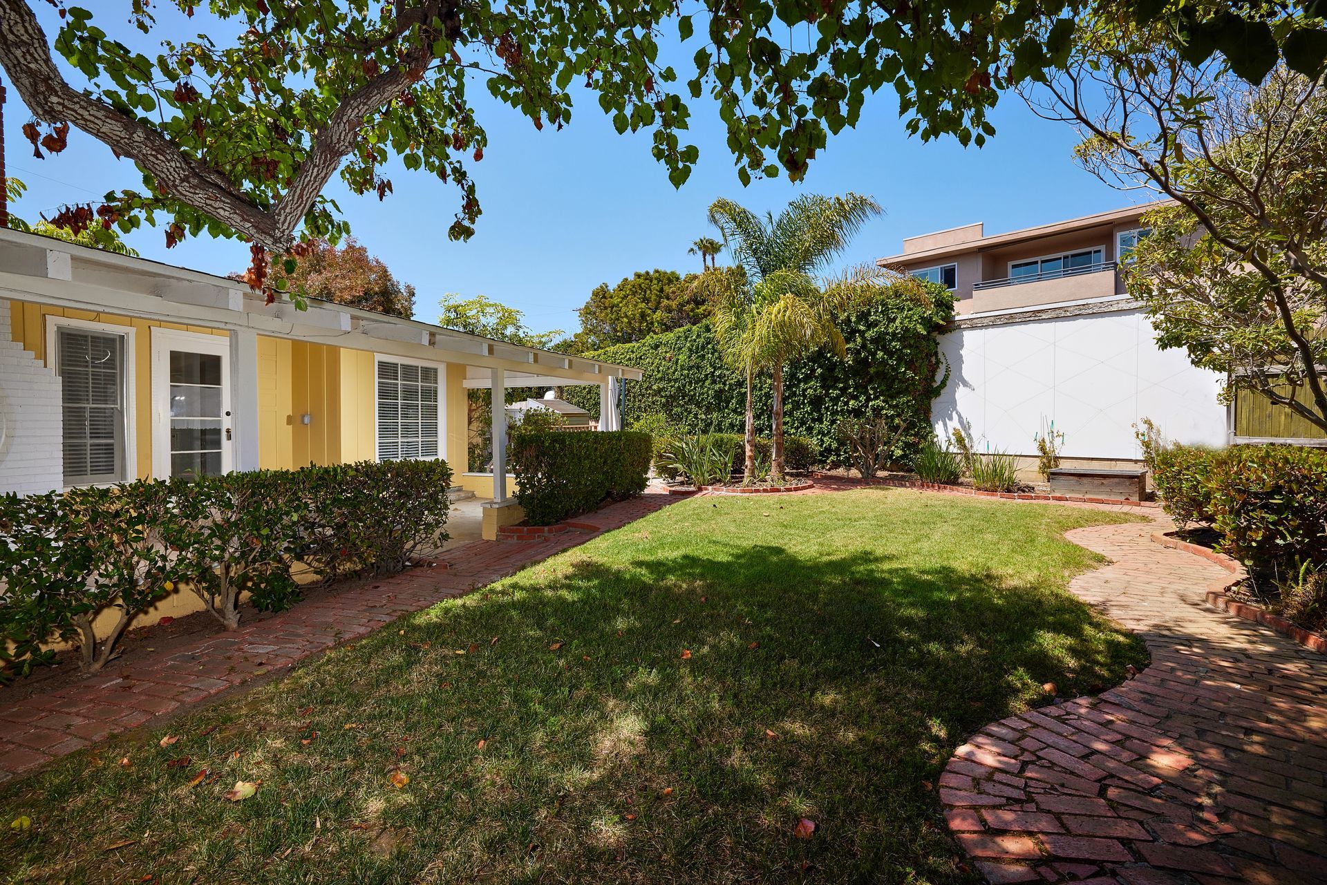 Sunny backyard courtyard with green lawn, brick path, and white and yellow houses