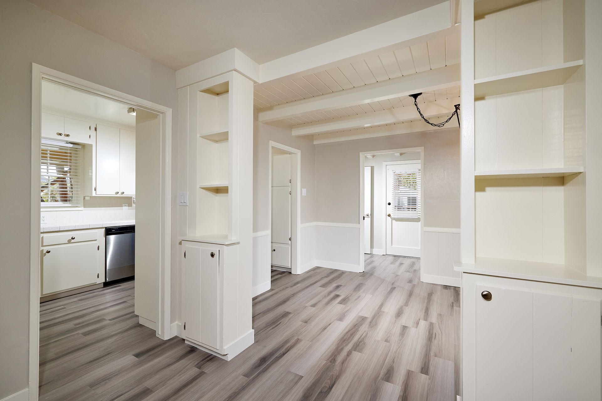Bright empty open-plan room with built-in shelves, wood floors, and a view into the kitchen