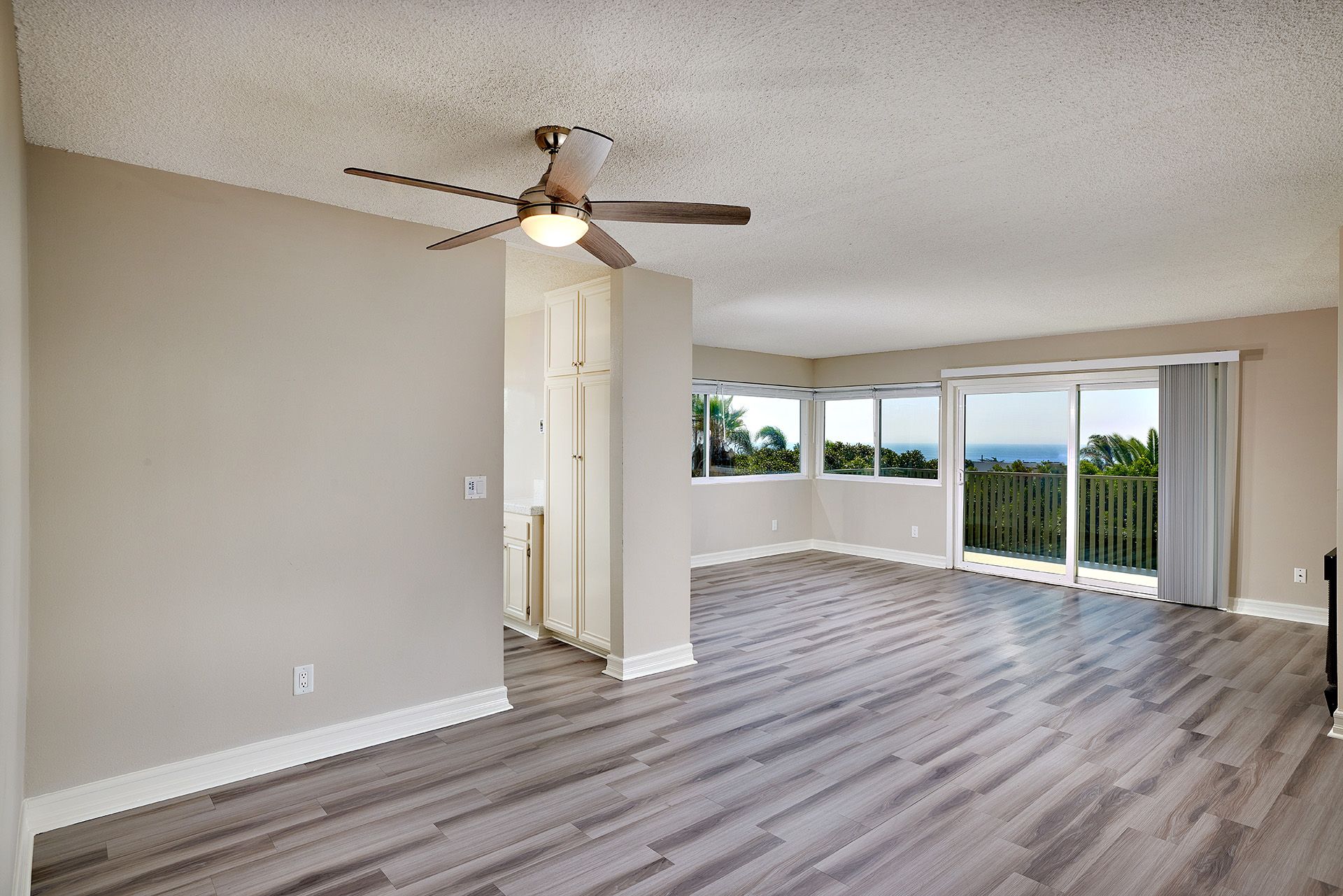 Empty living room with gray flooring, ceiling fan, and large windows overlooking greenery.