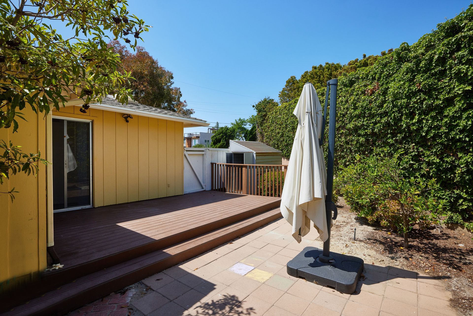 Patio with yellow house wall, wooden deck, umbrella stand, and green trees in bright sunlight