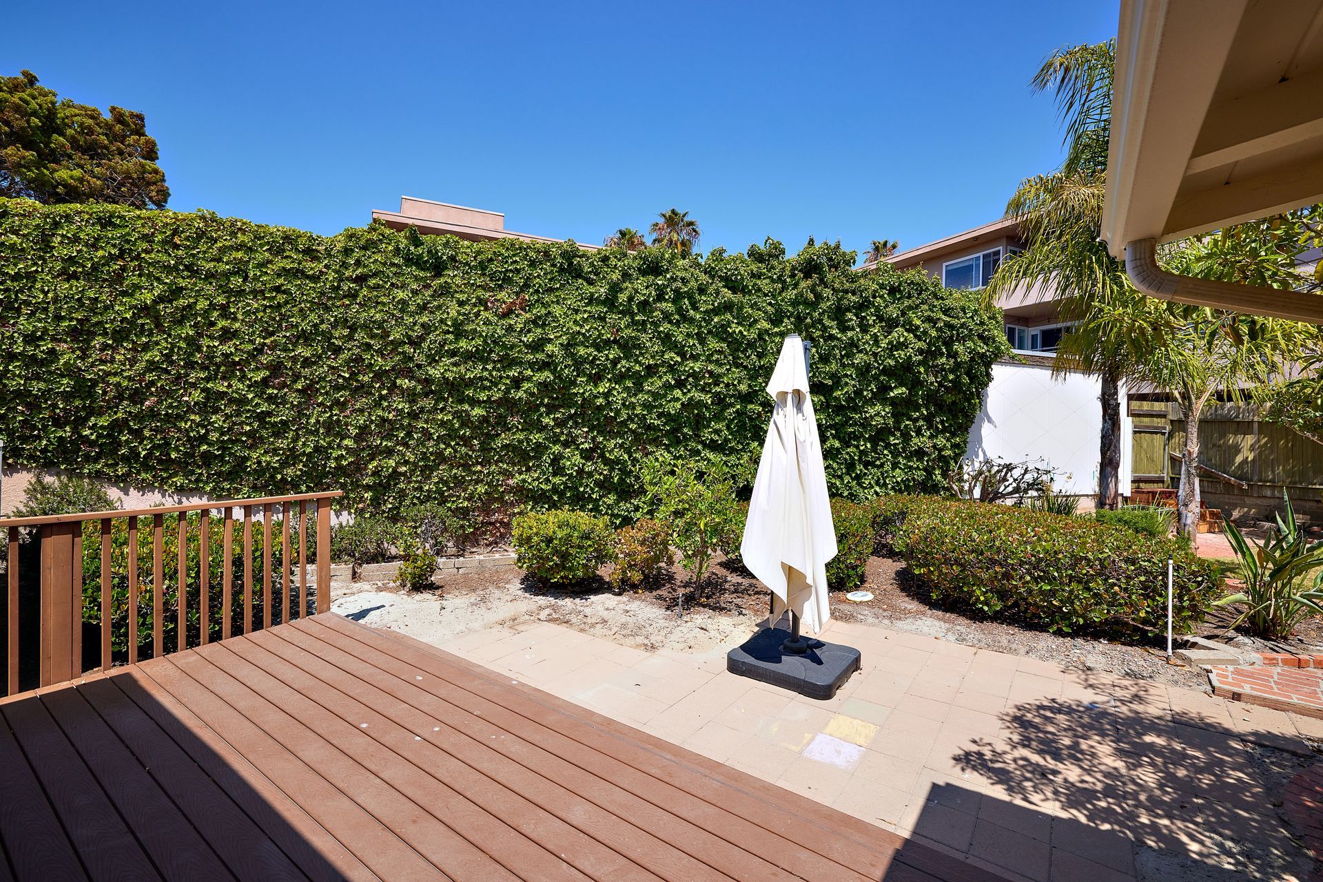 Patio with wooden deck, closed white umbrella, hedge fence, and sunny garden seating area