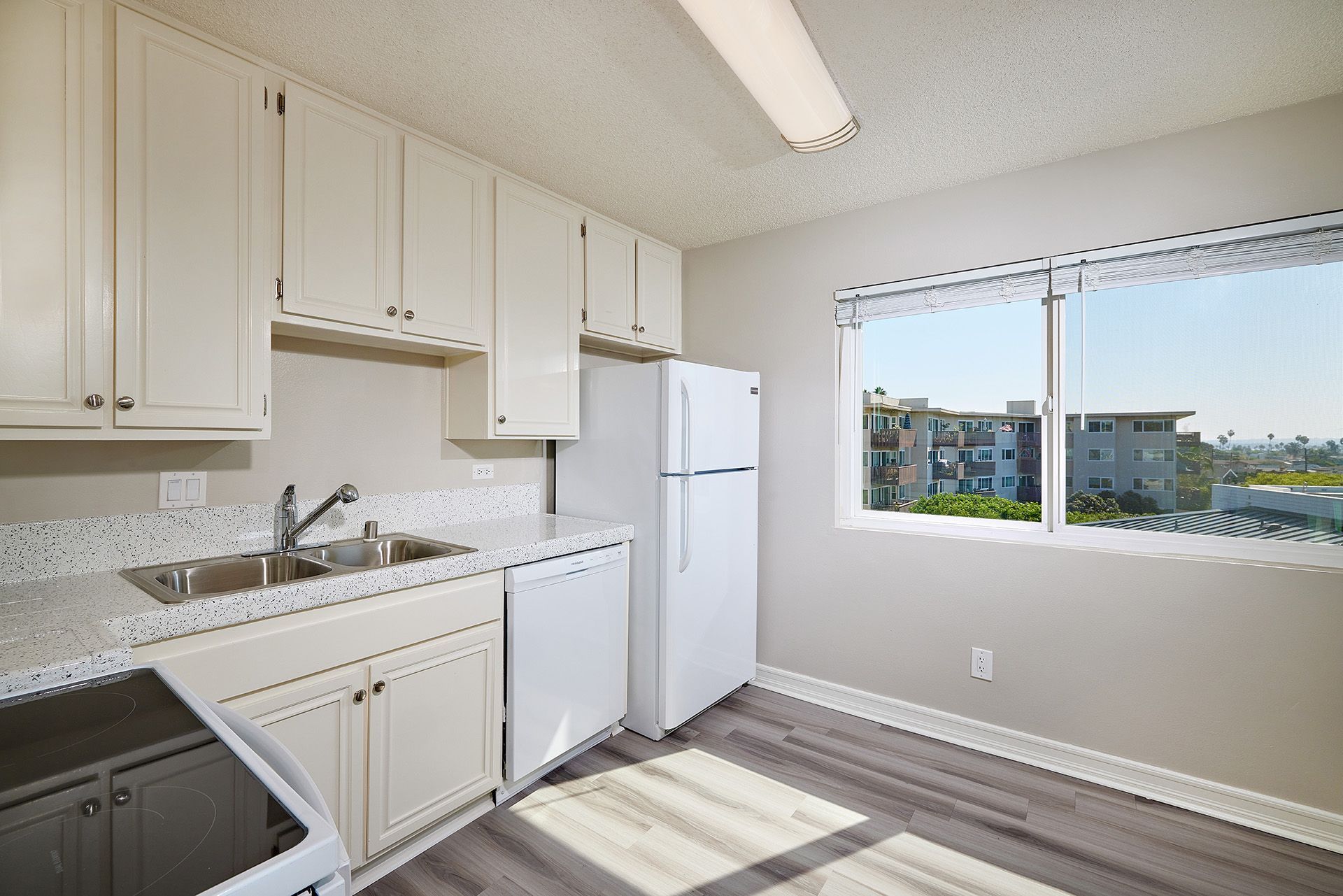 Bright kitchen with white cabinets, stove, sink, refrigerator, and a large window overlooking buildings.