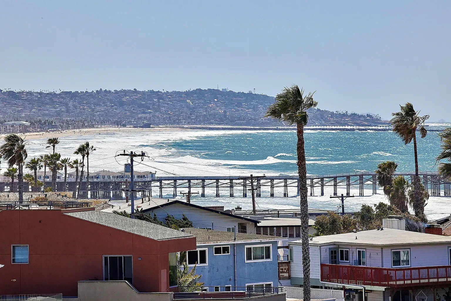 Coastal town with palm trees, waterfront buildings, and ocean waves under a clear sky.