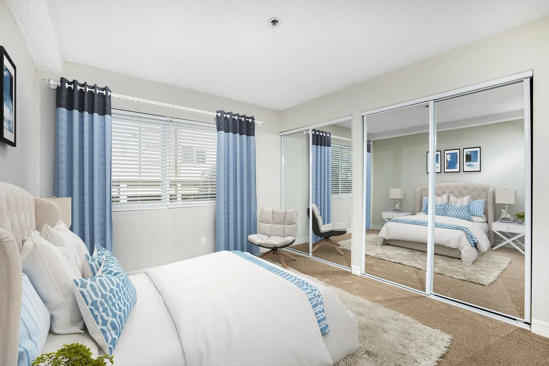 Bright bedroom with white bedding, blue curtains, mirrored closet doors, and a sitting chair near the window