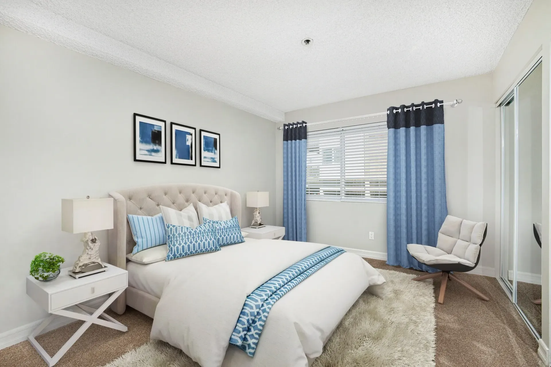 Bright bedroom with a blue-accented bed, nightstands, window curtains, and a white chair.