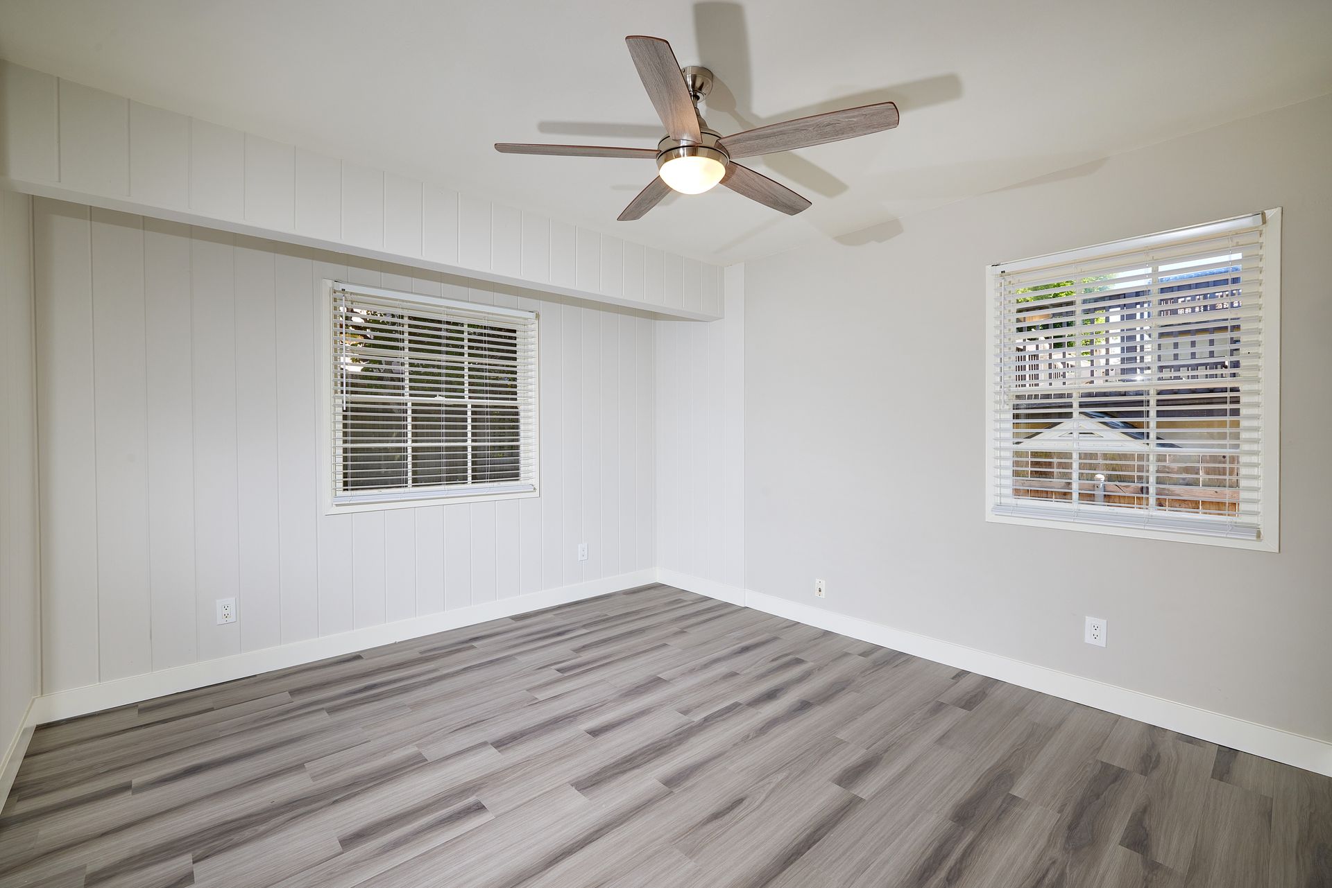 Empty white bedroom with gray wood flooring, ceiling fan, and two windows with blinds