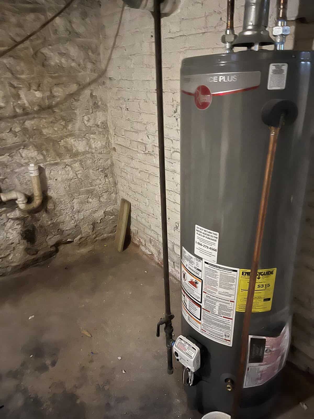 Water heater in a basement. The tank is gray, next to a brick wall. A long black pipe extends down.