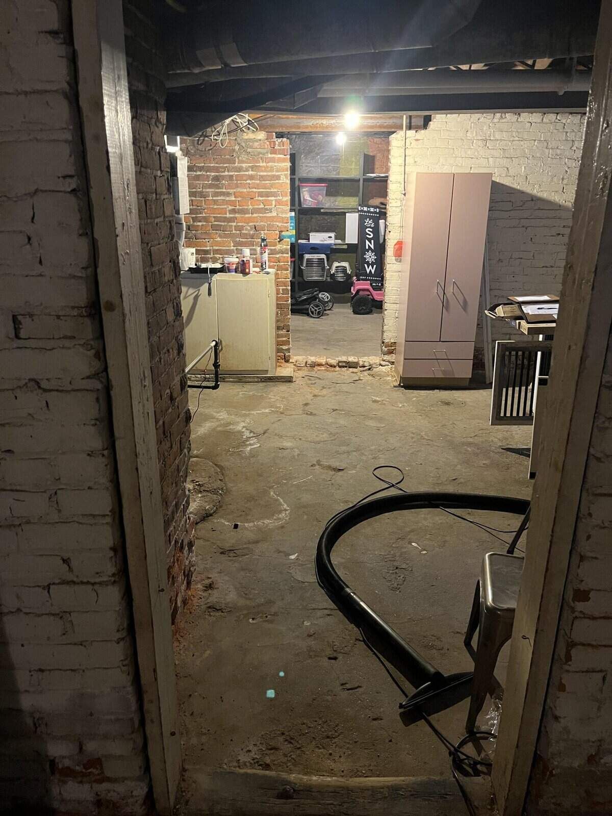 View through an interior doorway: unfinished basement with exposed brick, concrete floor, and storage.