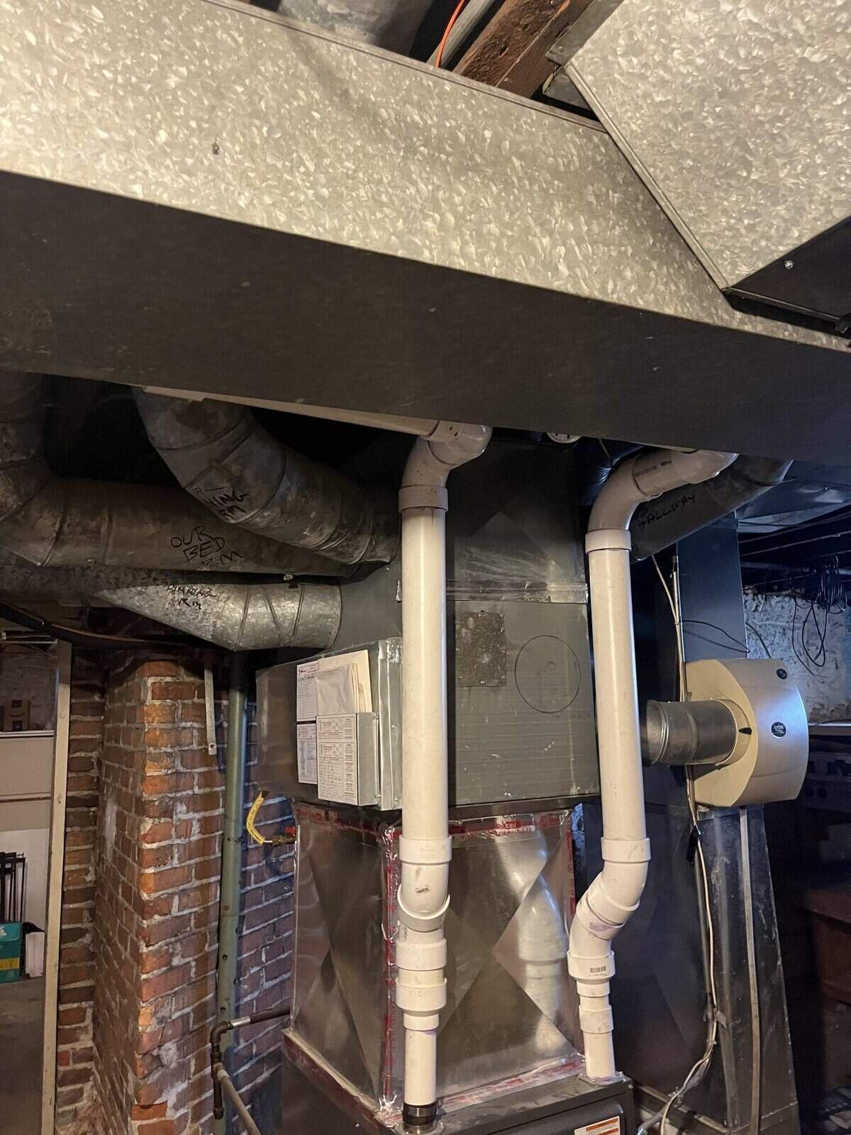 HVAC unit in a basement. Ductwork and white PVC pipes are connected. Brick wall on the left.
