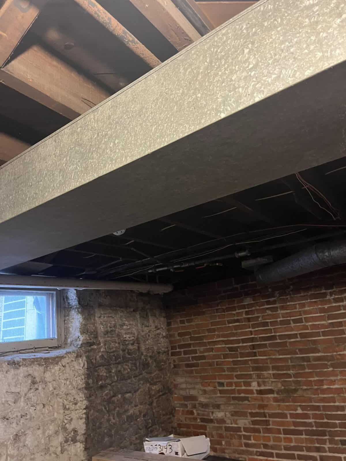 Basement interior with metal beam, brick wall, window, and exposed ceiling joists.