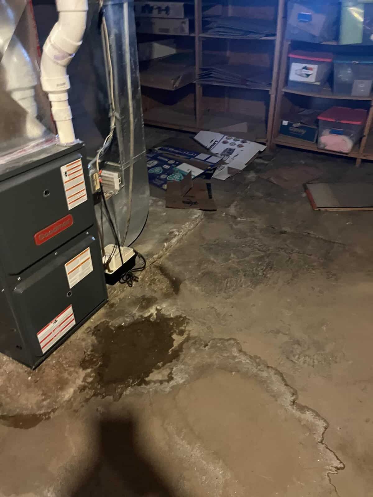 Basement with furnace and water damage on concrete floor. Shelving with boxes in the background.