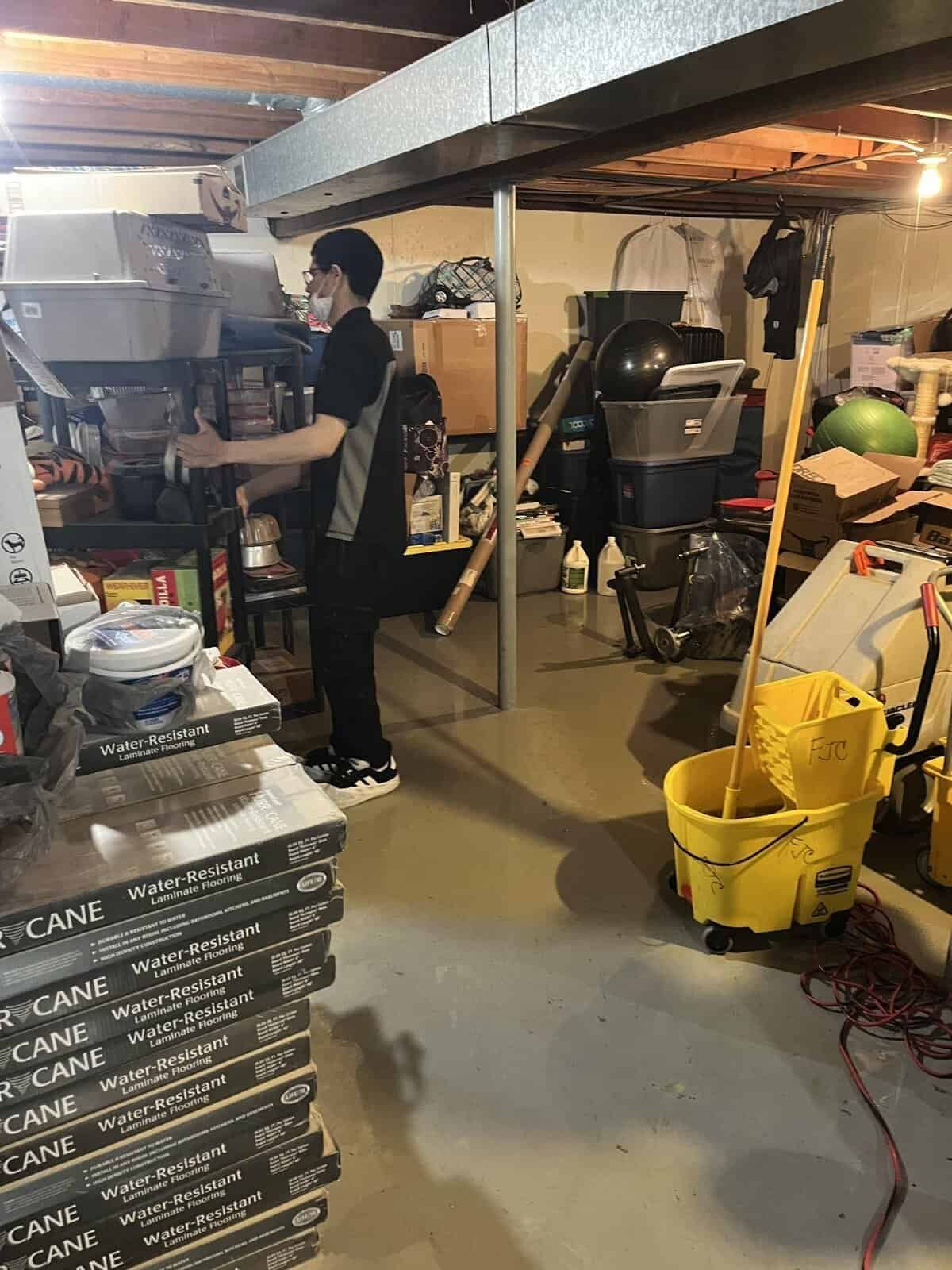 Person in a basement stands near stacked flooring and a mop bucket; cluttered shelves and support beams are visible.