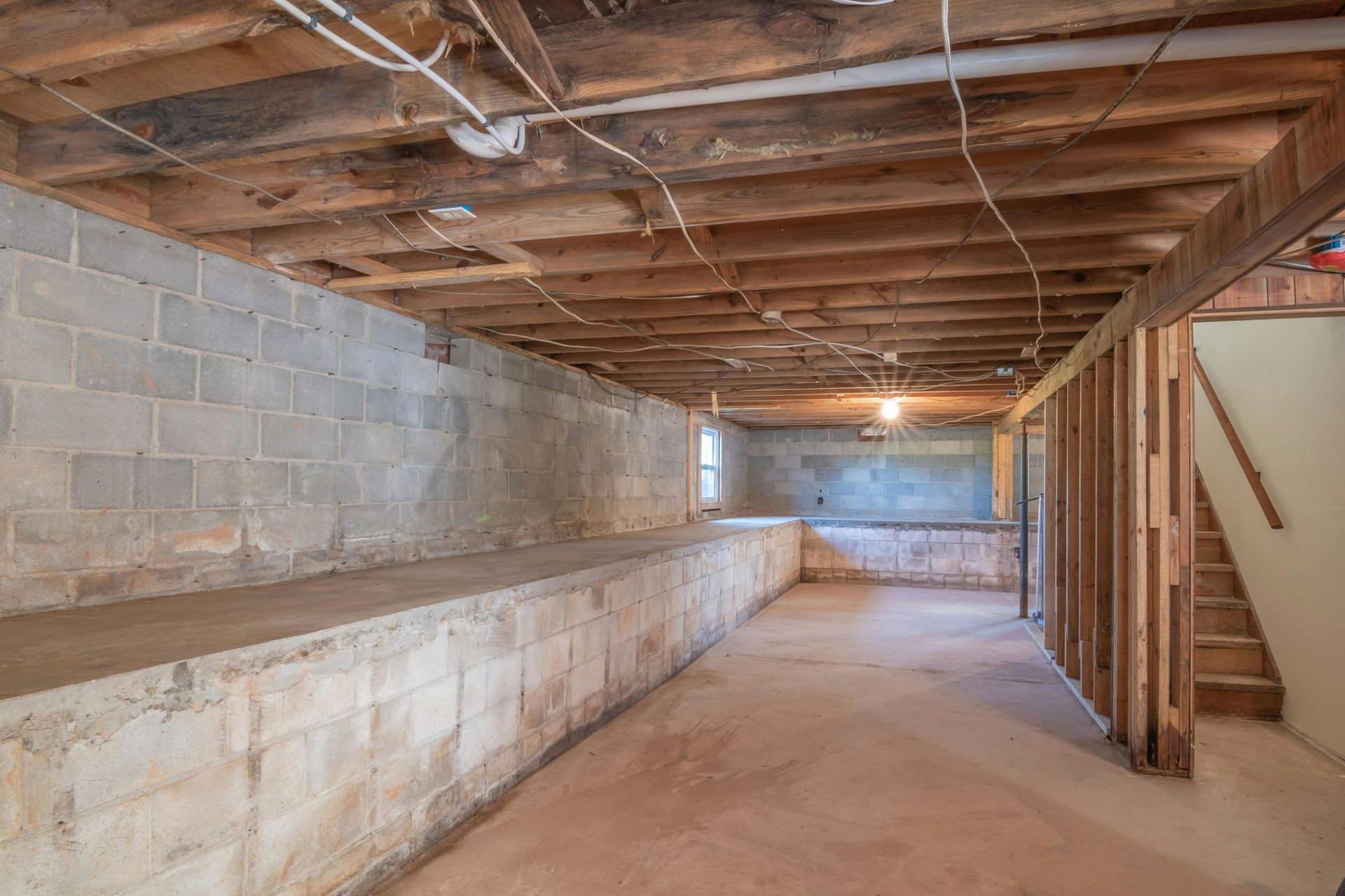 Empty basement with concrete block walls, exposed wooden beams, and a concrete floor.