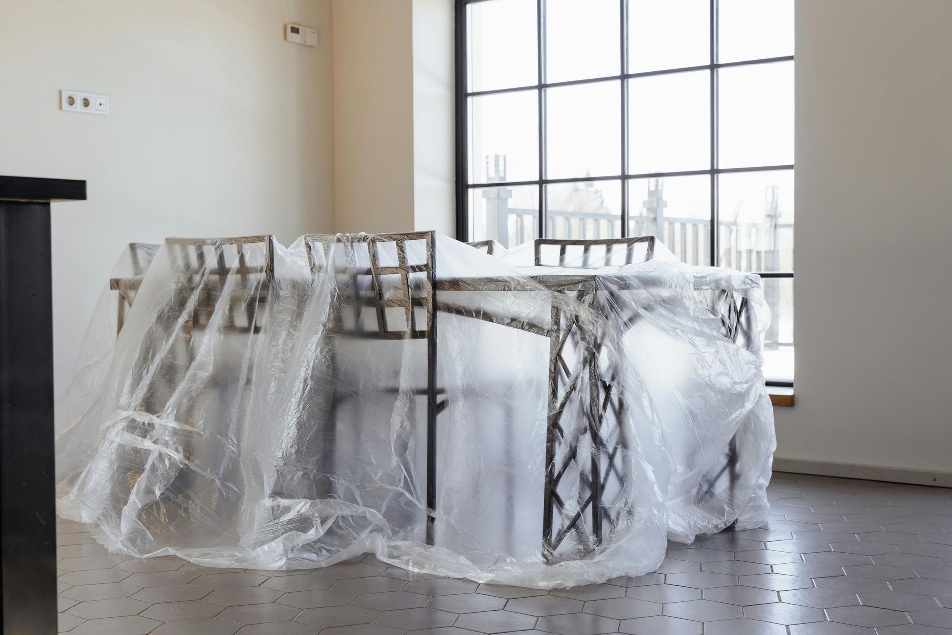 Dining table and chairs covered in clear plastic sheeting, in a bright room with large window.