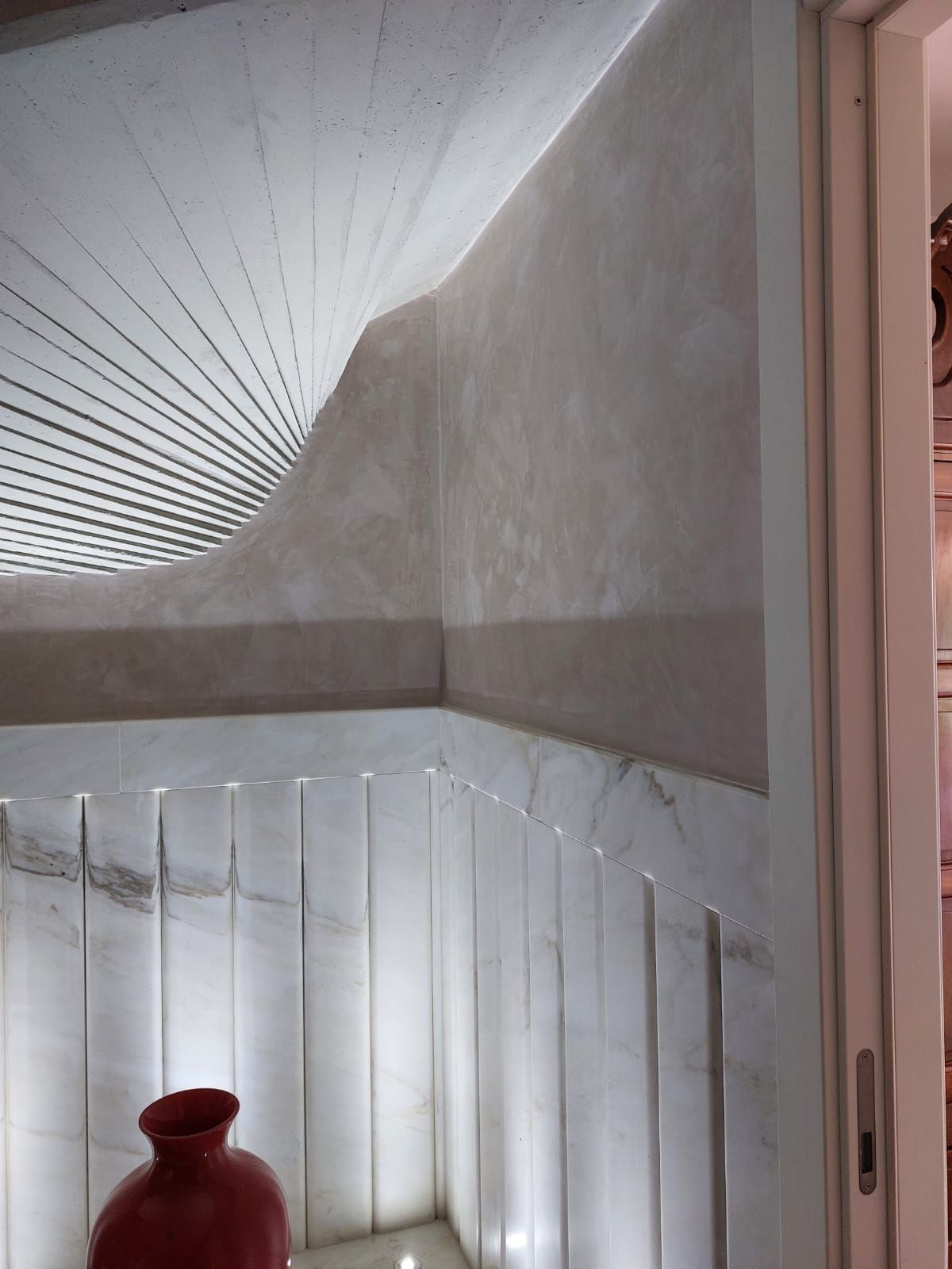 Un angolo interno moderno con pareti bianche testurizzate, un soffitto decorato e accenti verticali in marmo illuminati da luci da incasso, un vaso rosso.