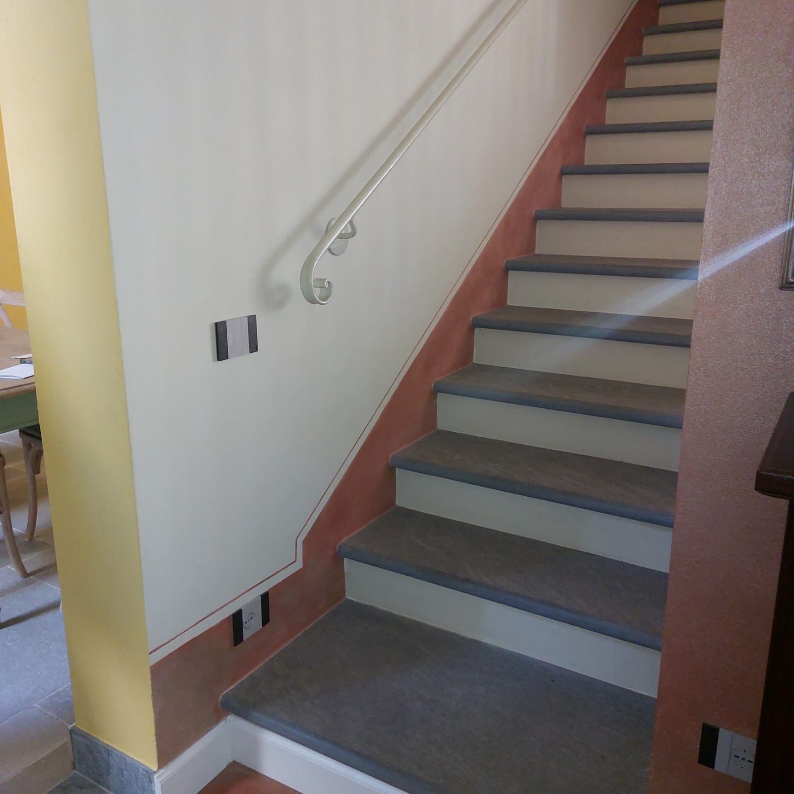 Scala con gradini grigi, finiture rosse e corrimano argentato su pareti color crema e giallo.