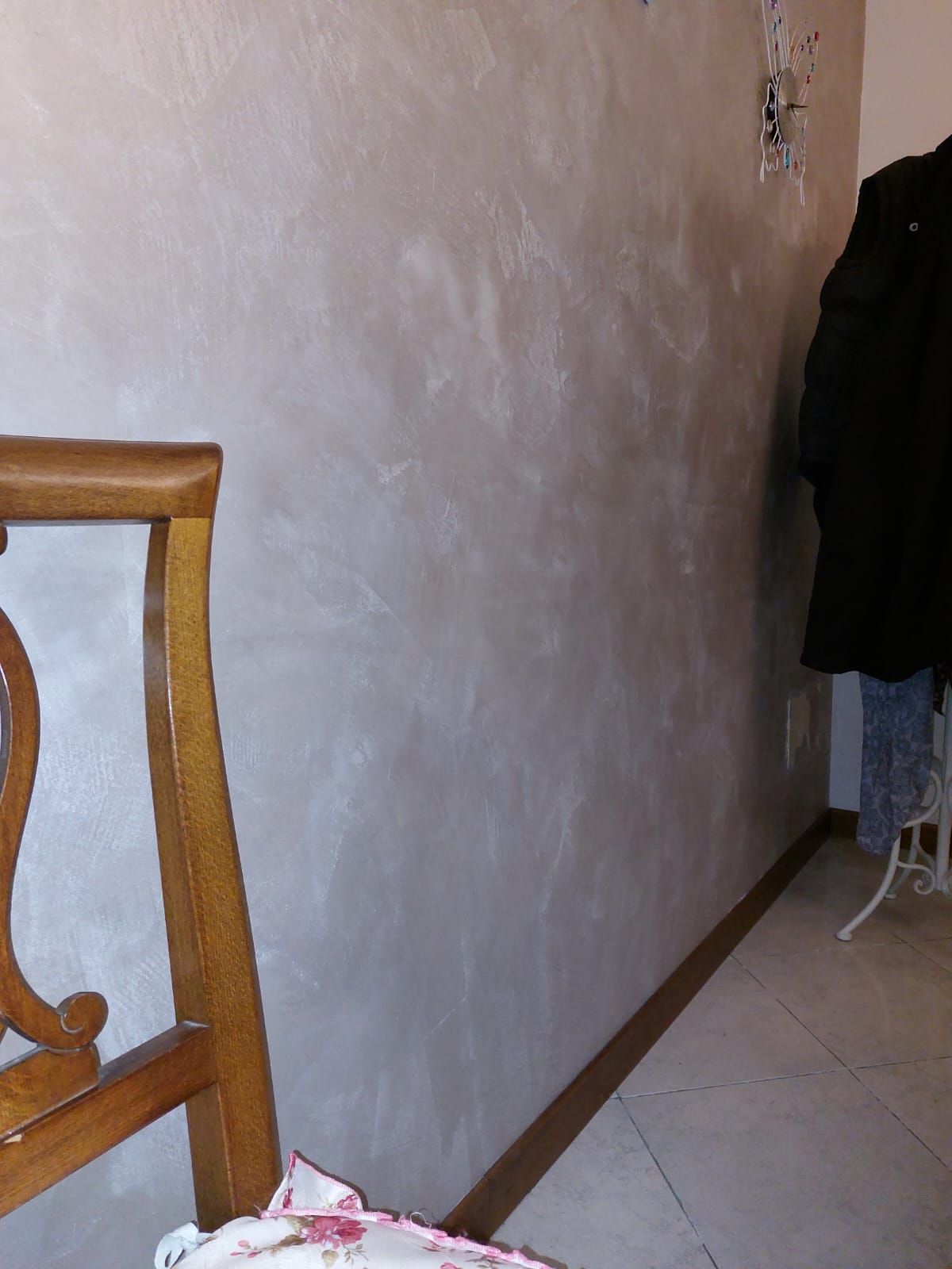Una sedia di legno vicino a una parete testurizzata e di colore chiaro con uno zoccolo marrone scuro. Cappotti appesi su un attaccapanni sullo sfondo.