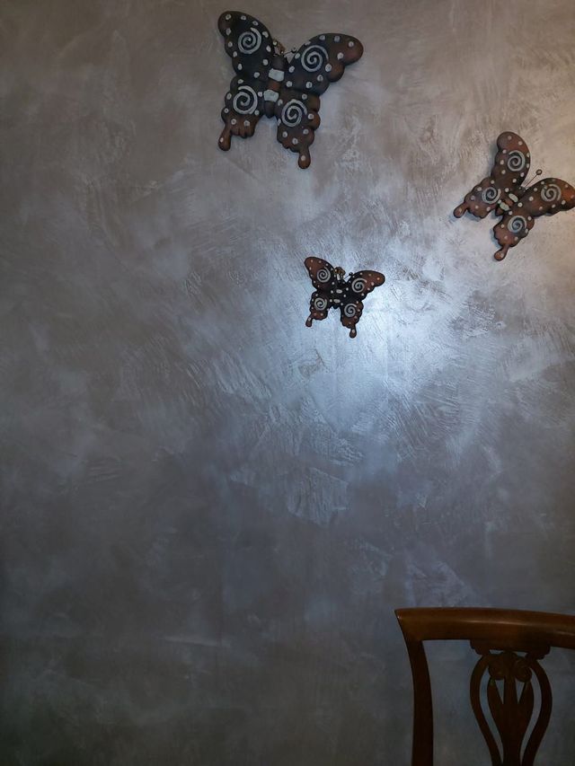 Tre arazzi decorativi a forma di farfalla su una parete ruvida dall'aspetto metallico. È visibile una parte di una sedia in legno.