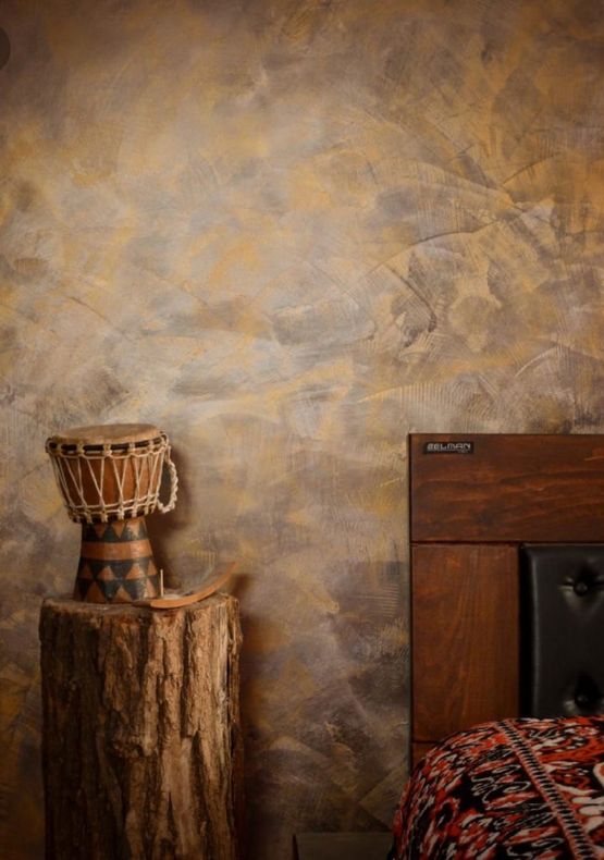 Un tamburo djembe è appoggiato su un ceppo d'albero accanto a una testiera in legno scuro. La parete retrostante è decorata con texture marrone e oro.