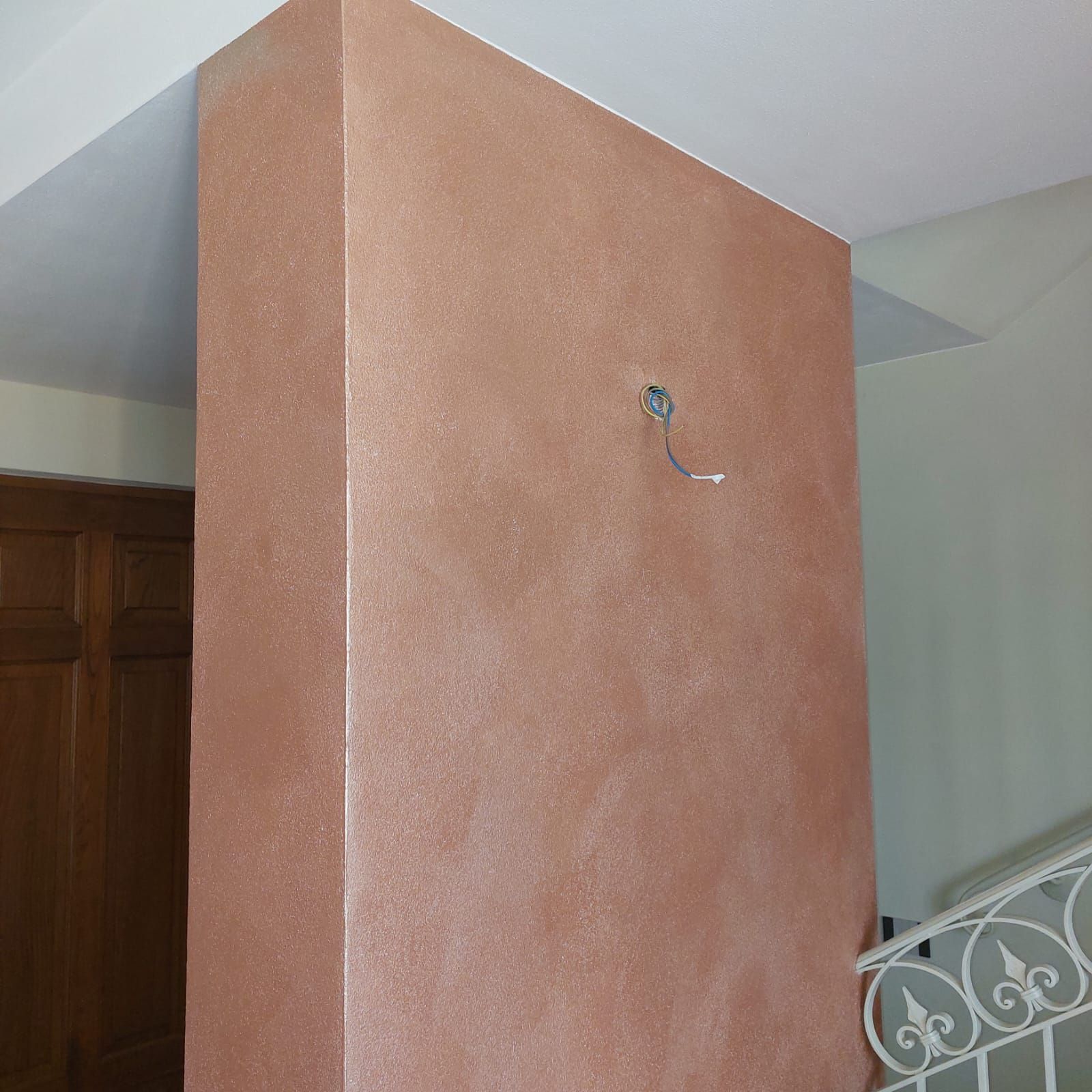 Un muro ruvido color rame. Un filo metallico nudo pende dal muro. Nelle vicinanze si vedono una scala e una porta di legno.