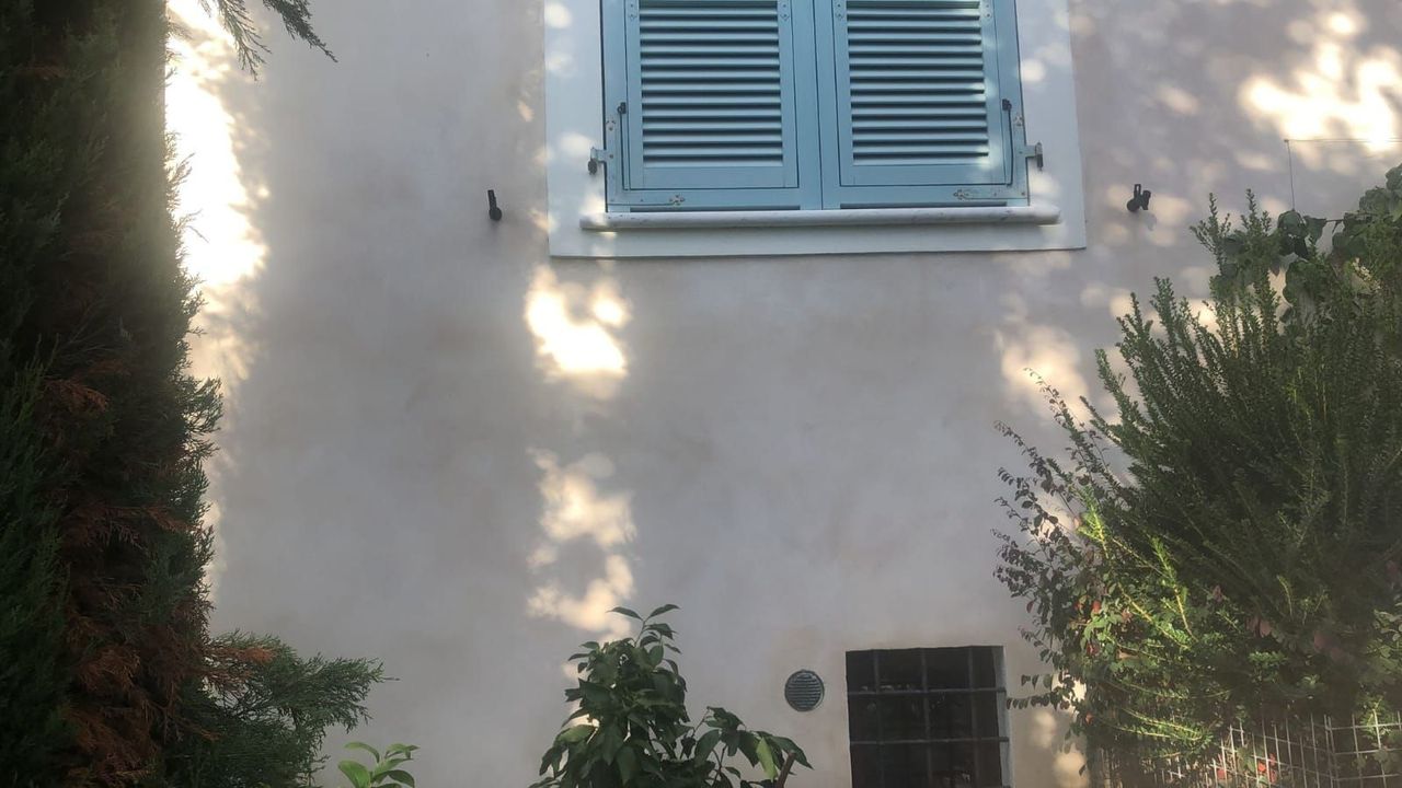 Un edificio di colore chiaro con una finestra con persiane blu, circondato da alberi che proiettano ombre.