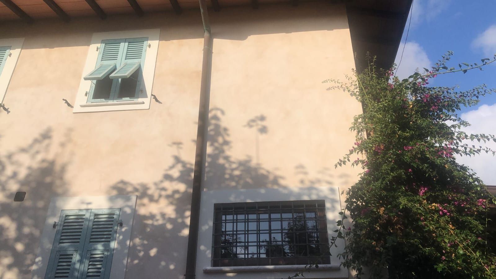 Edificio in stucco beige con persiane azzurre e una finestra scura. Una pianta rampicante con fiori rossi cresce lungo il lato destro.