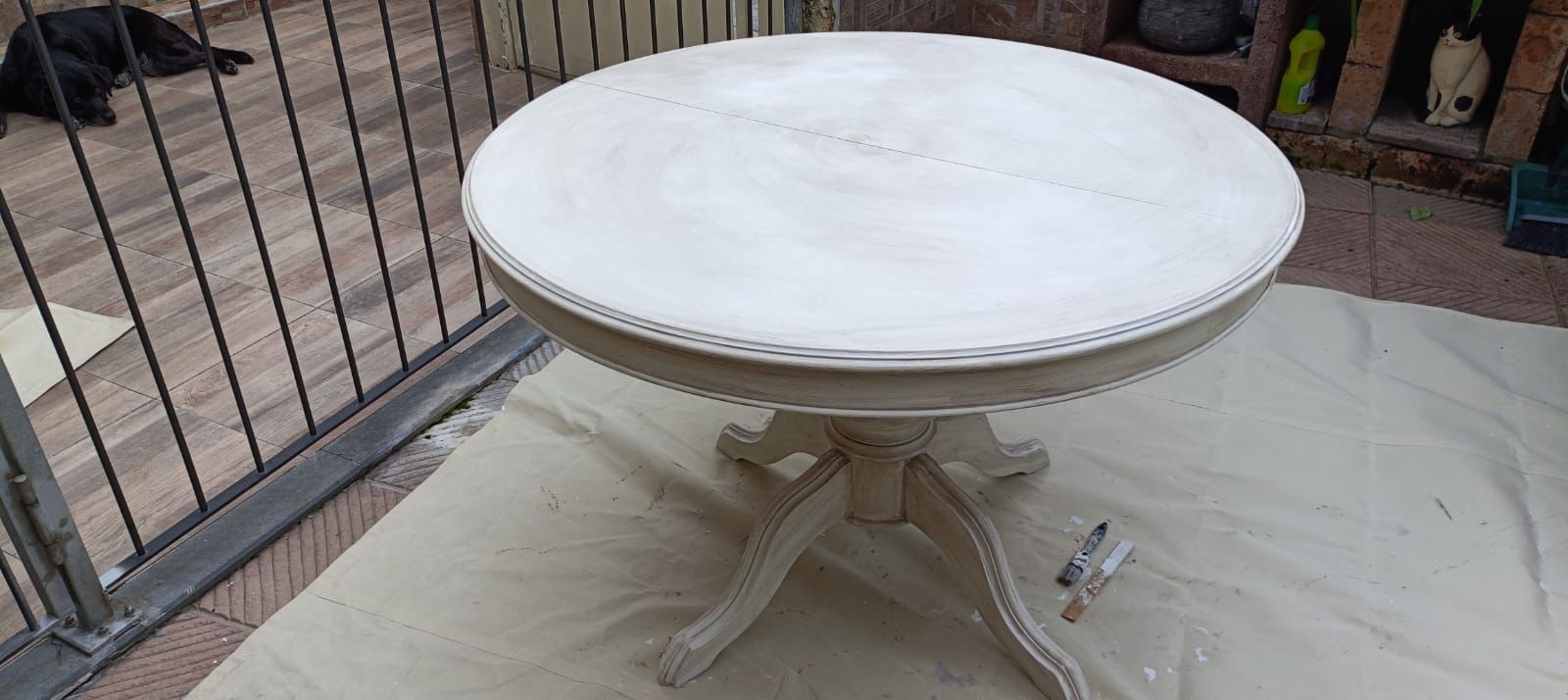 Un tavolo rotondo dipinto di bianco con gambe decorate è appoggiato su un telo protettivo all'esterno. Sullo sfondo si vede un cane.