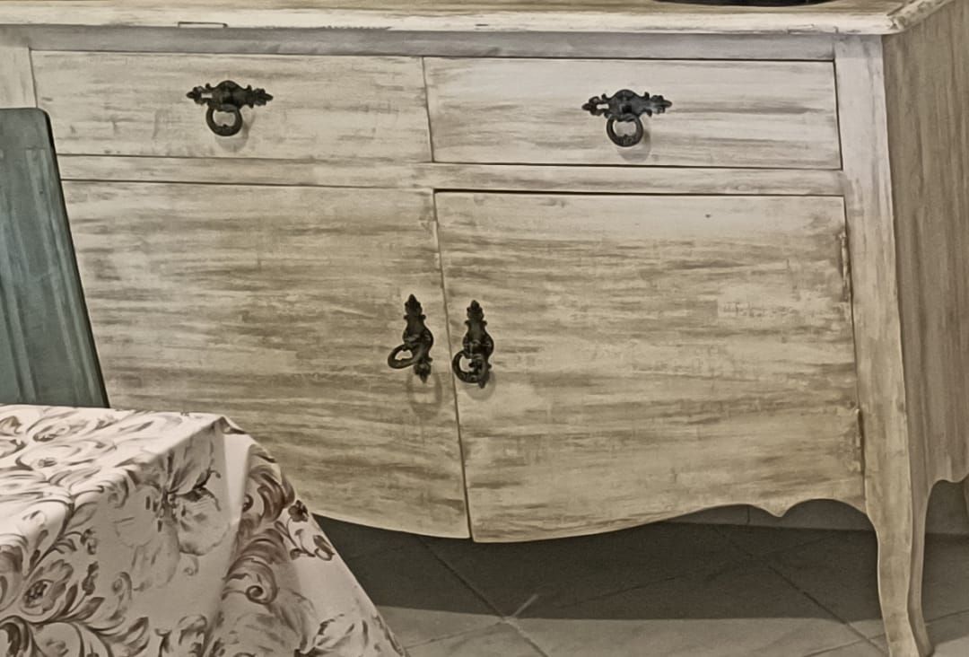 Mobiletto bianco anticato con due cassetti e due ante, maniglie in metallo scuro decorato. In primo piano un tavolo con una tovaglia floreale.