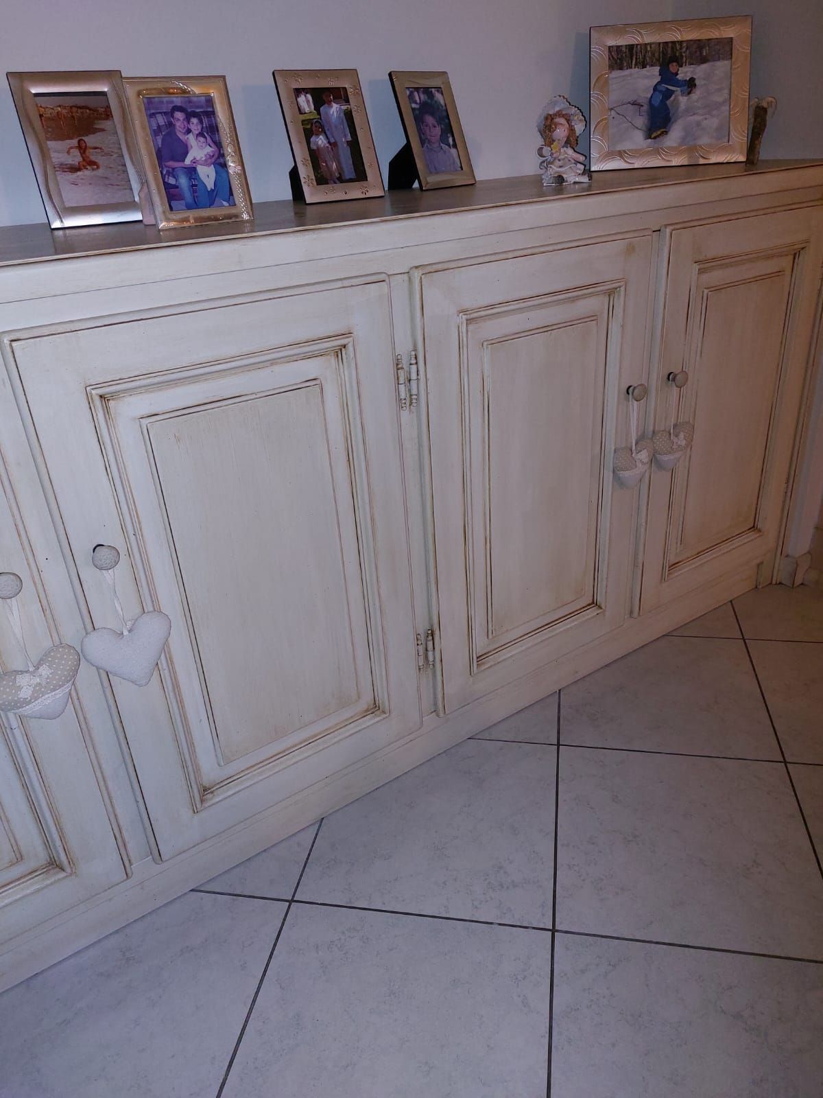 Mobile color crema con foto incorniciate sul piano, appoggiato a una parete bianca. Il mobile presenta maniglie decorate e una finitura anticata.