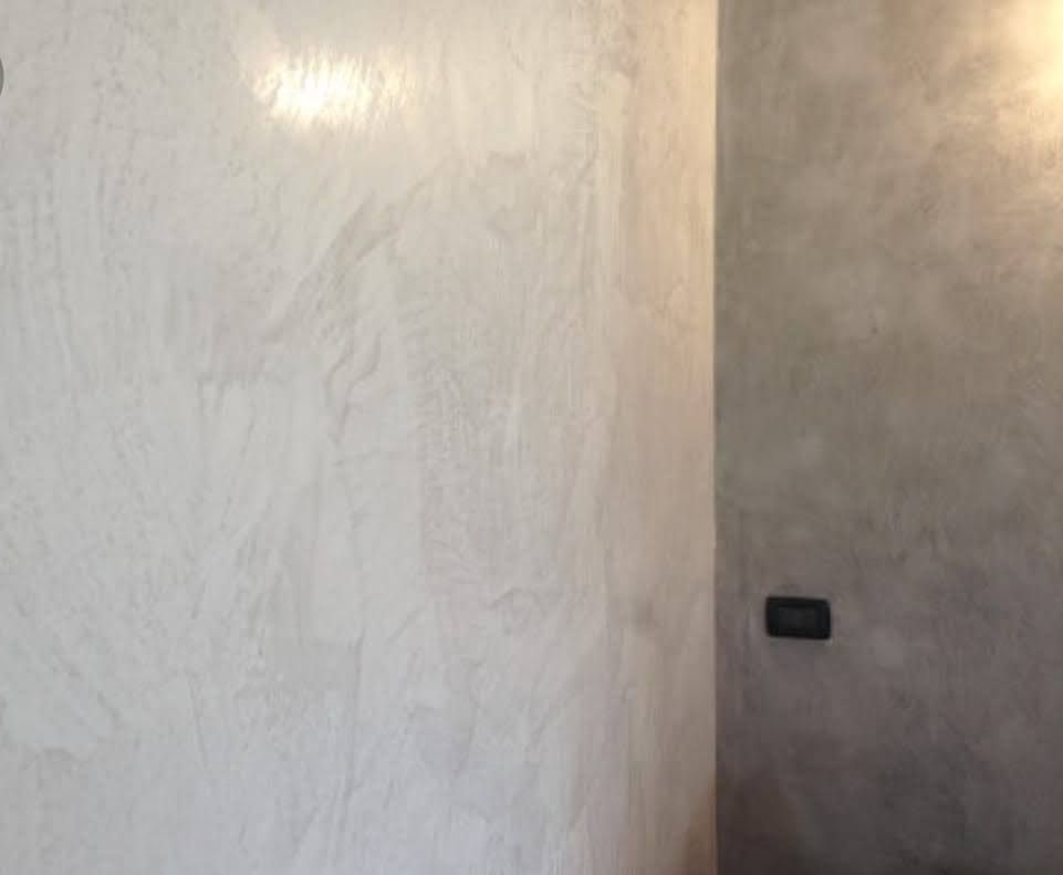 Due pareti adiacenti con intonaco chiaro e testurizzato. Una parete è prevalentemente bianca, l'altra grigia. Sulla parete grigia è visibile una placca nera per gli interruttori.