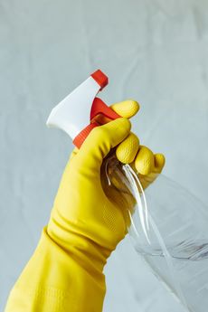 Hand in yellow glove holding a spray bottle, spraying liquid against a gray background.