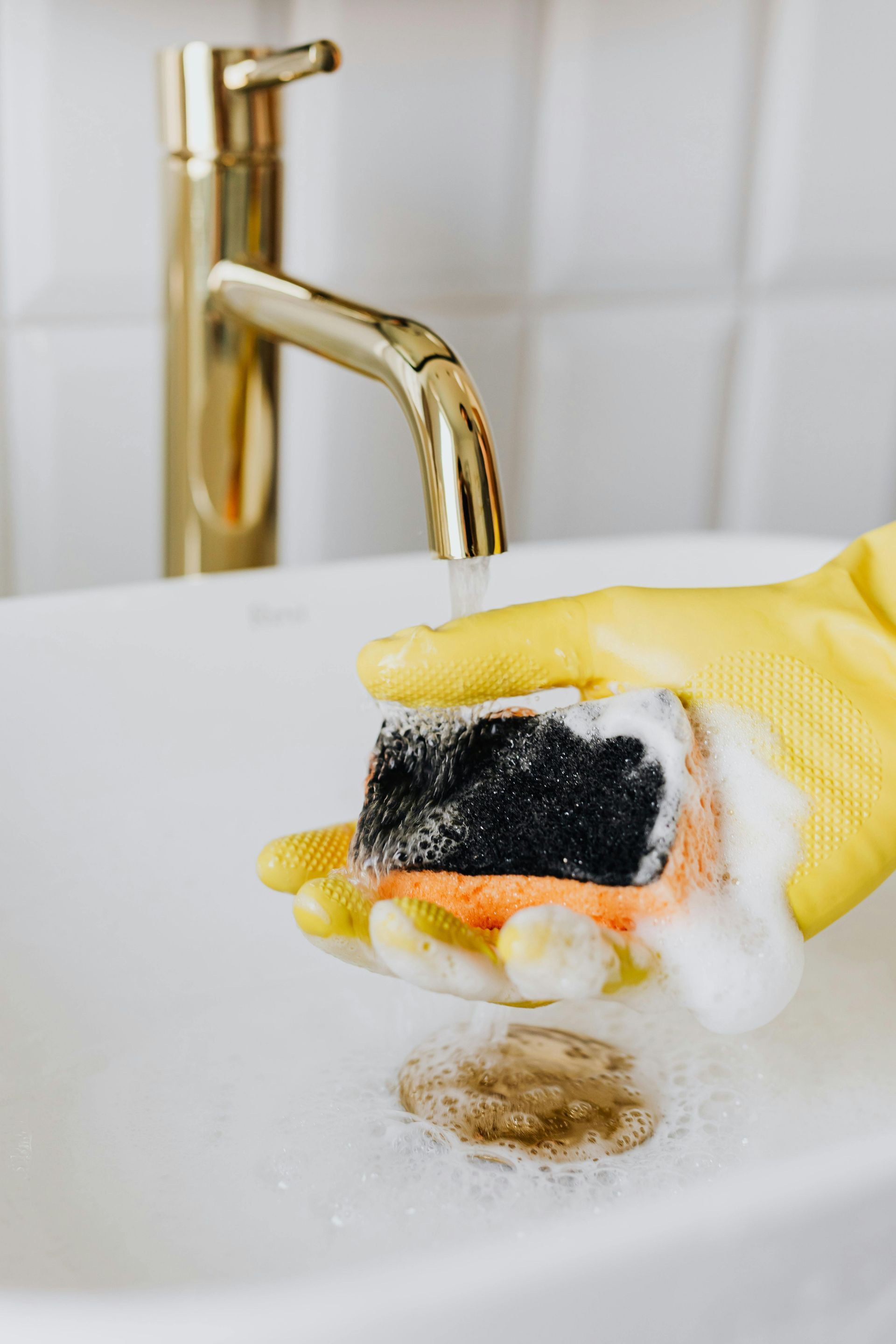 Hand in yellow glove scrubbing a white sink with a soapy sponge under a gold faucet.