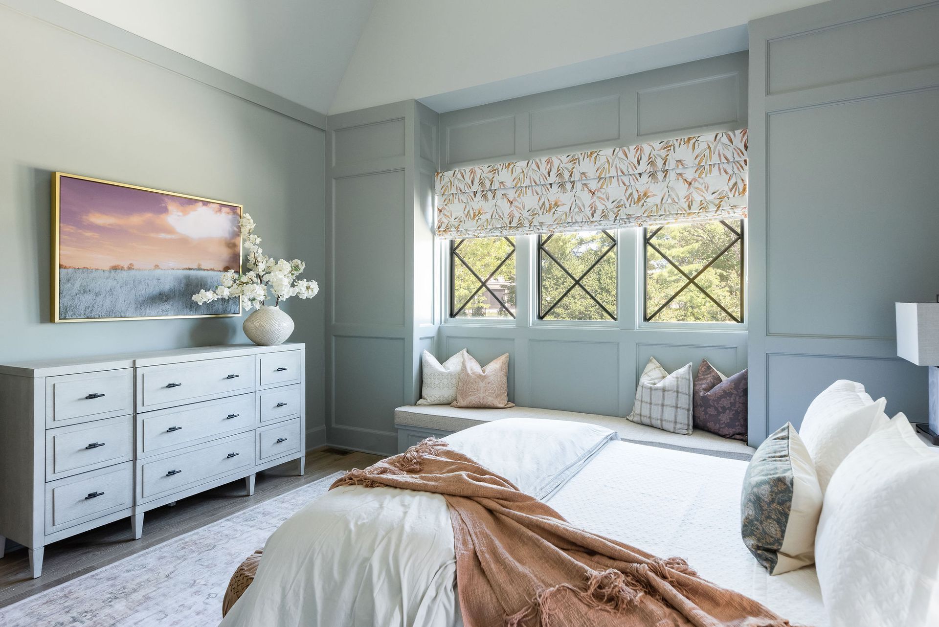Bright bedroom with light blue walls, white dresser, bed, floral window shade, and soft pink accents