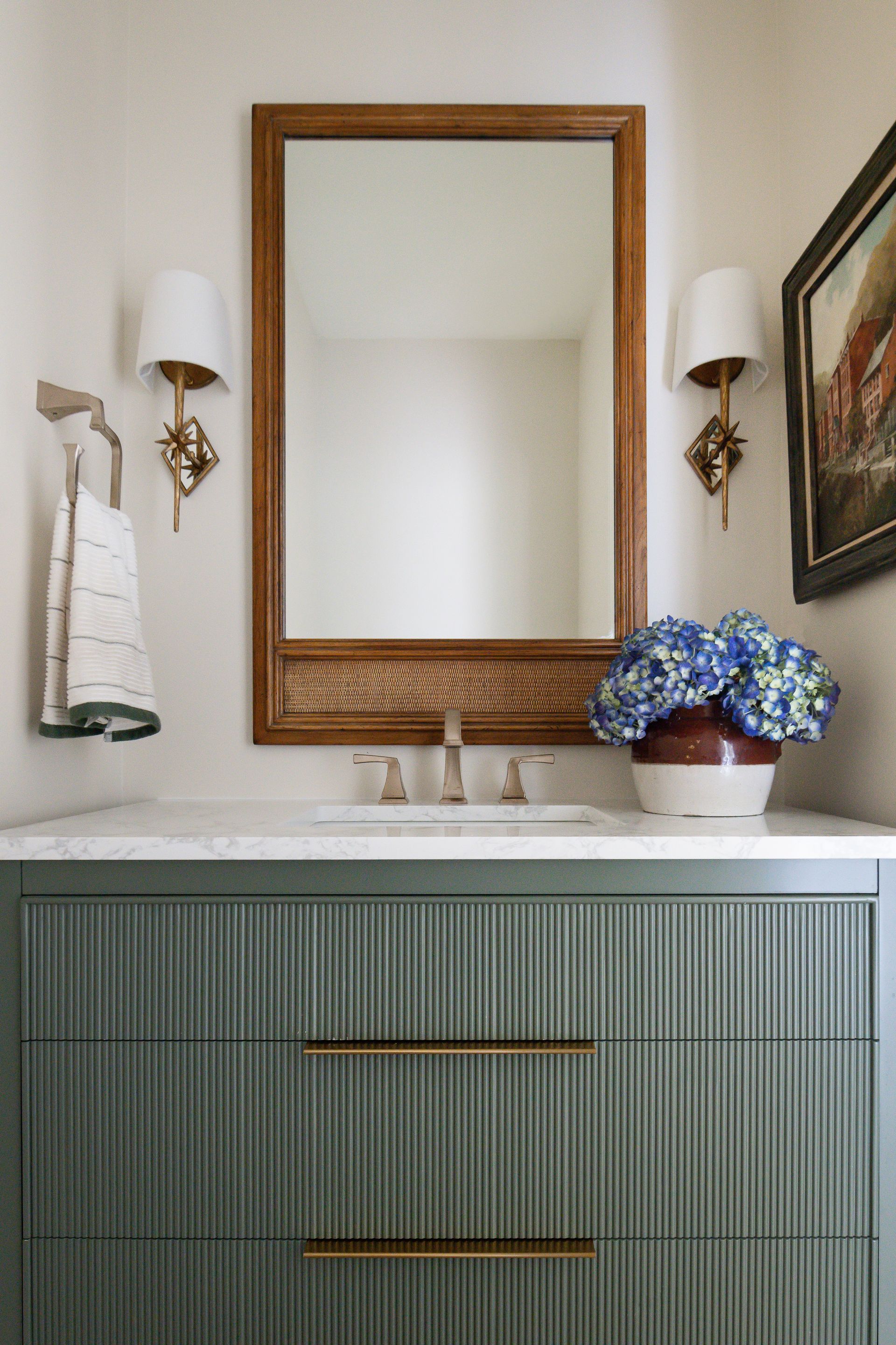 Green vanity with gold hardware, mirror, sconces, and blue flower arrangement.