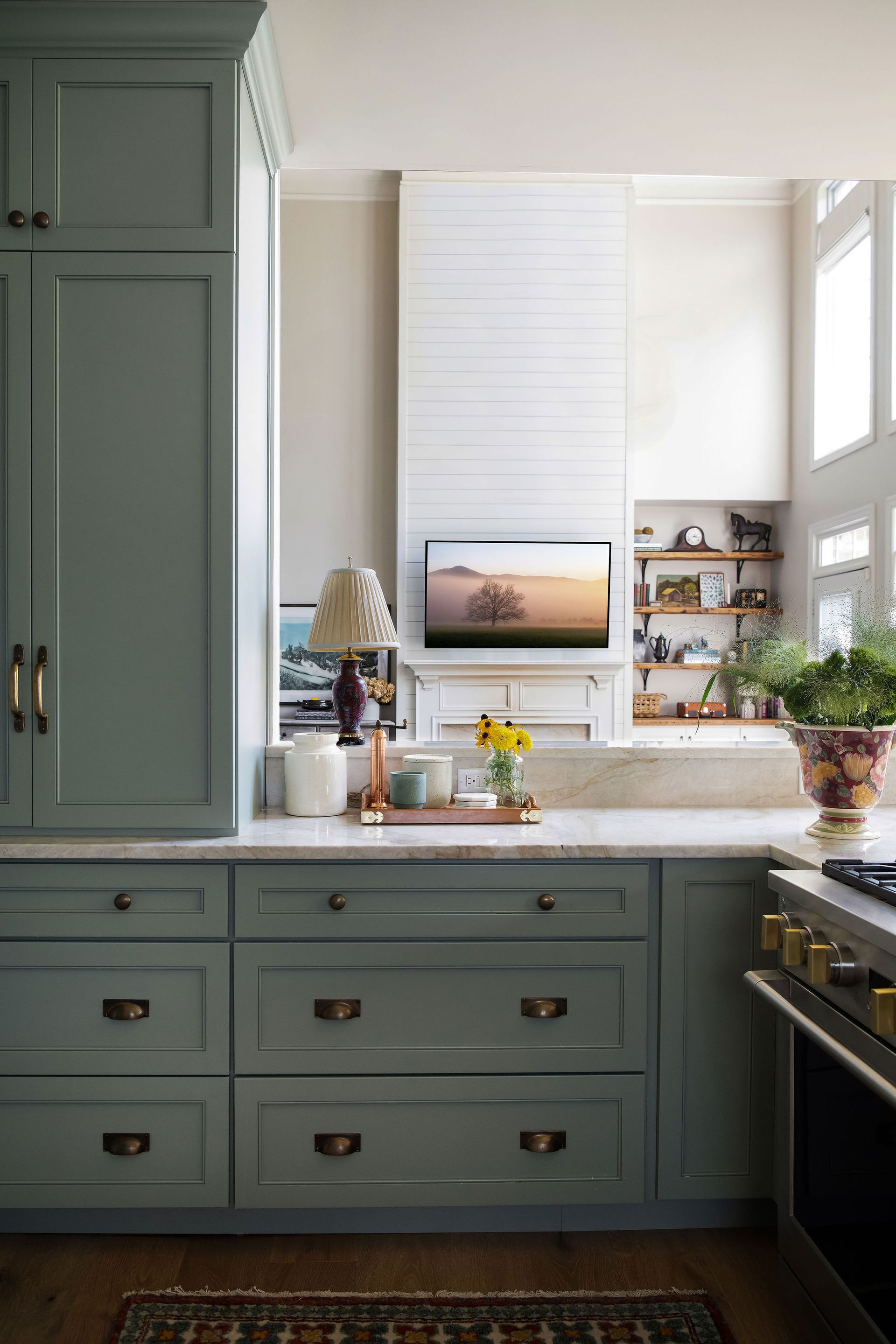 Green kitchen cabinets with marble countertop, TV above, and window with shelves.