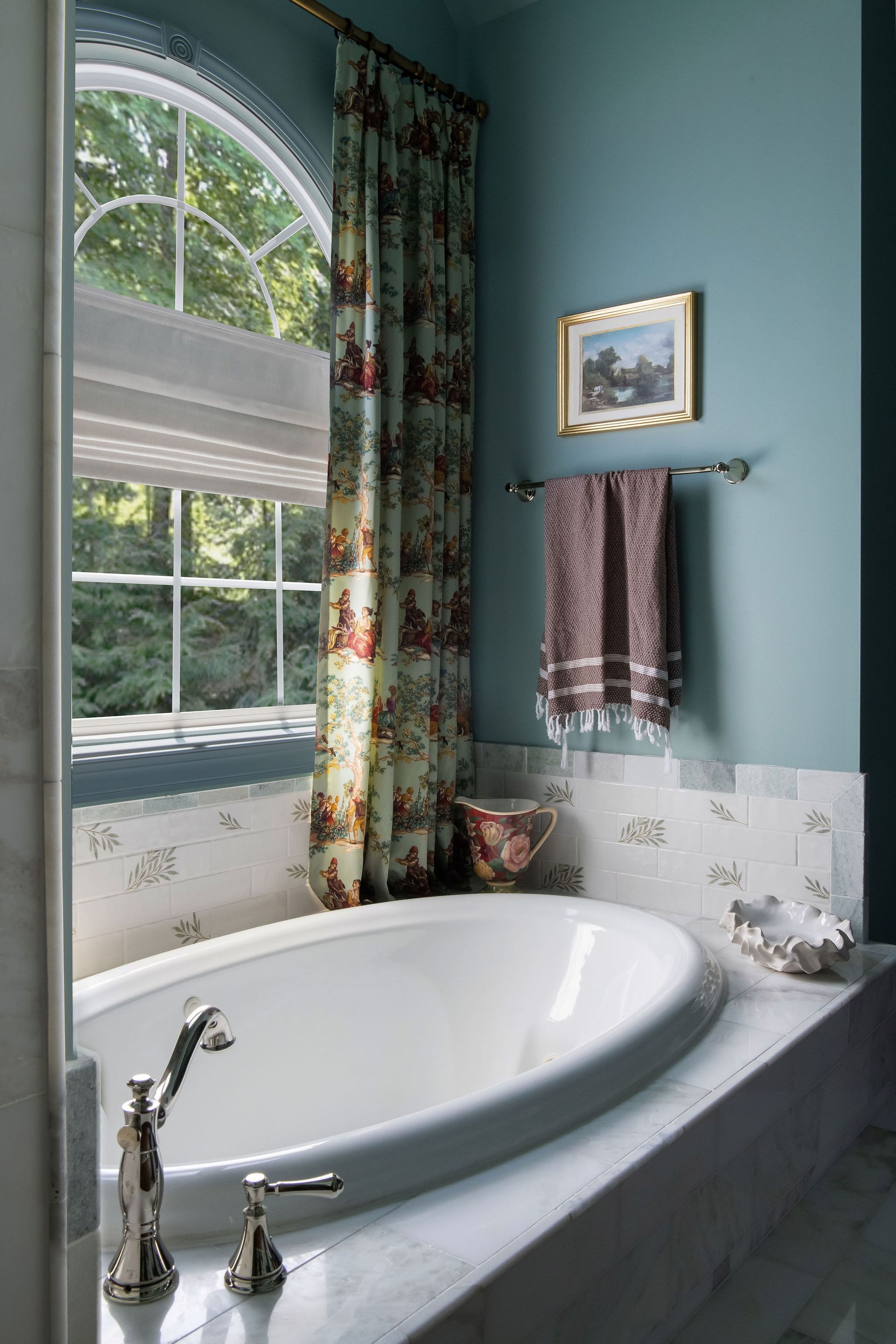 Bathroom with oval tub, teal wall, arched window, floral curtain, and artwork.