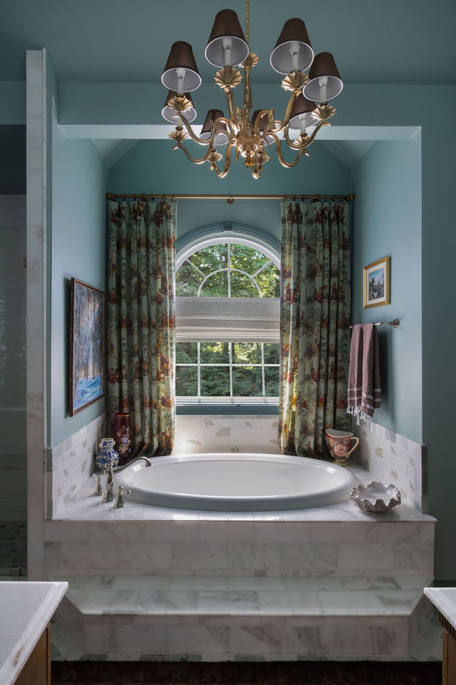A luxurious bathroom with an oval bathtub, floral curtains, a chandelier, and a large window overlooking trees.