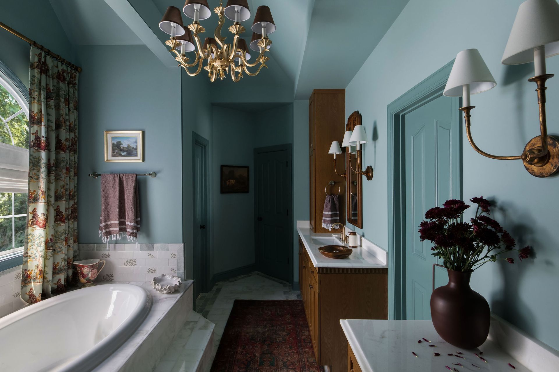 Blue bathroom with a bathtub, vanity, and a floral curtain. Gold chandelier and sconces.