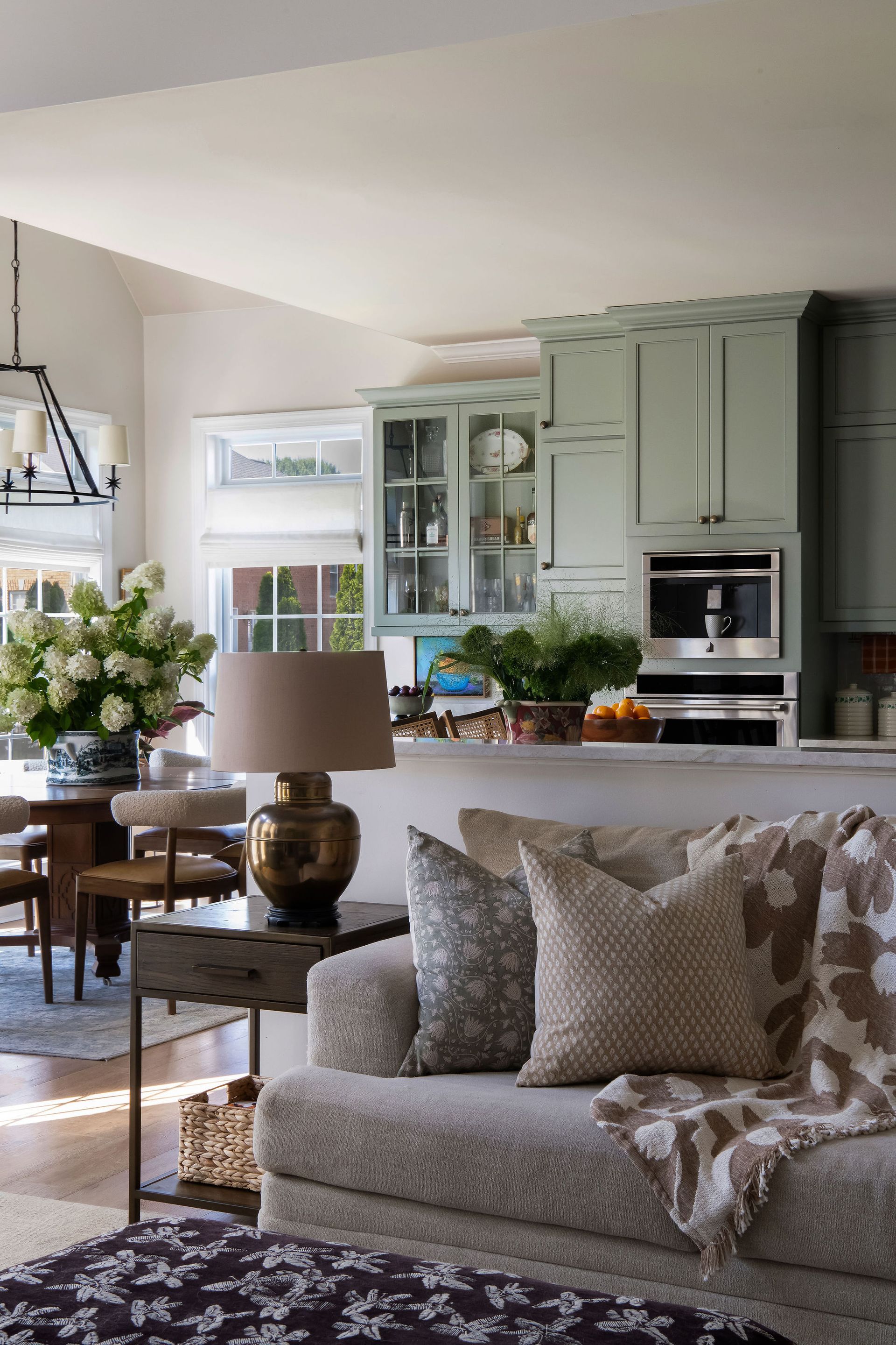Living room with light green kitchen. Sofa with textured pillows and blanket. Table lamp lit. Hydrangeas.