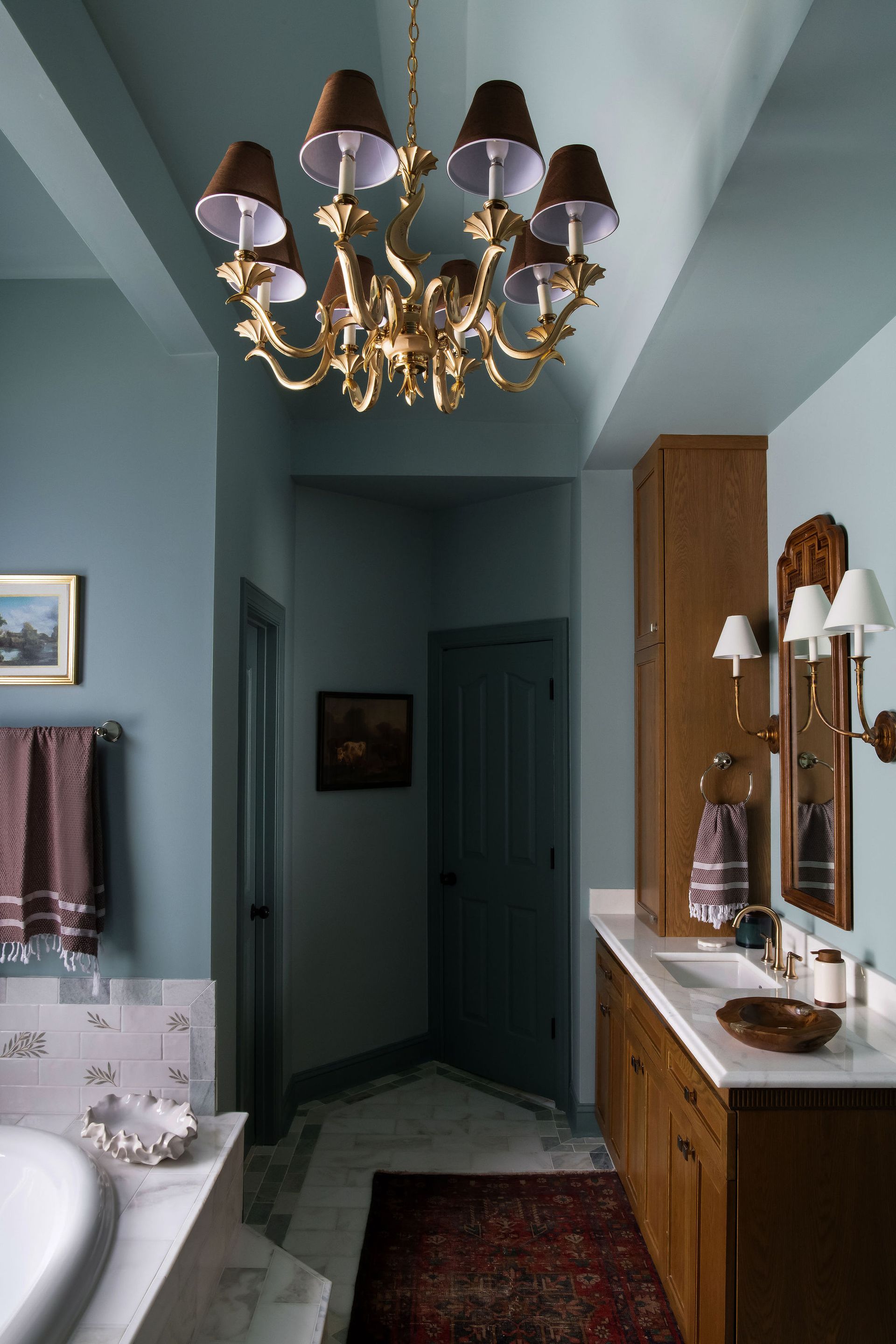 Bathroom with a gold chandelier, blue walls, wooden vanity, and a red rug.