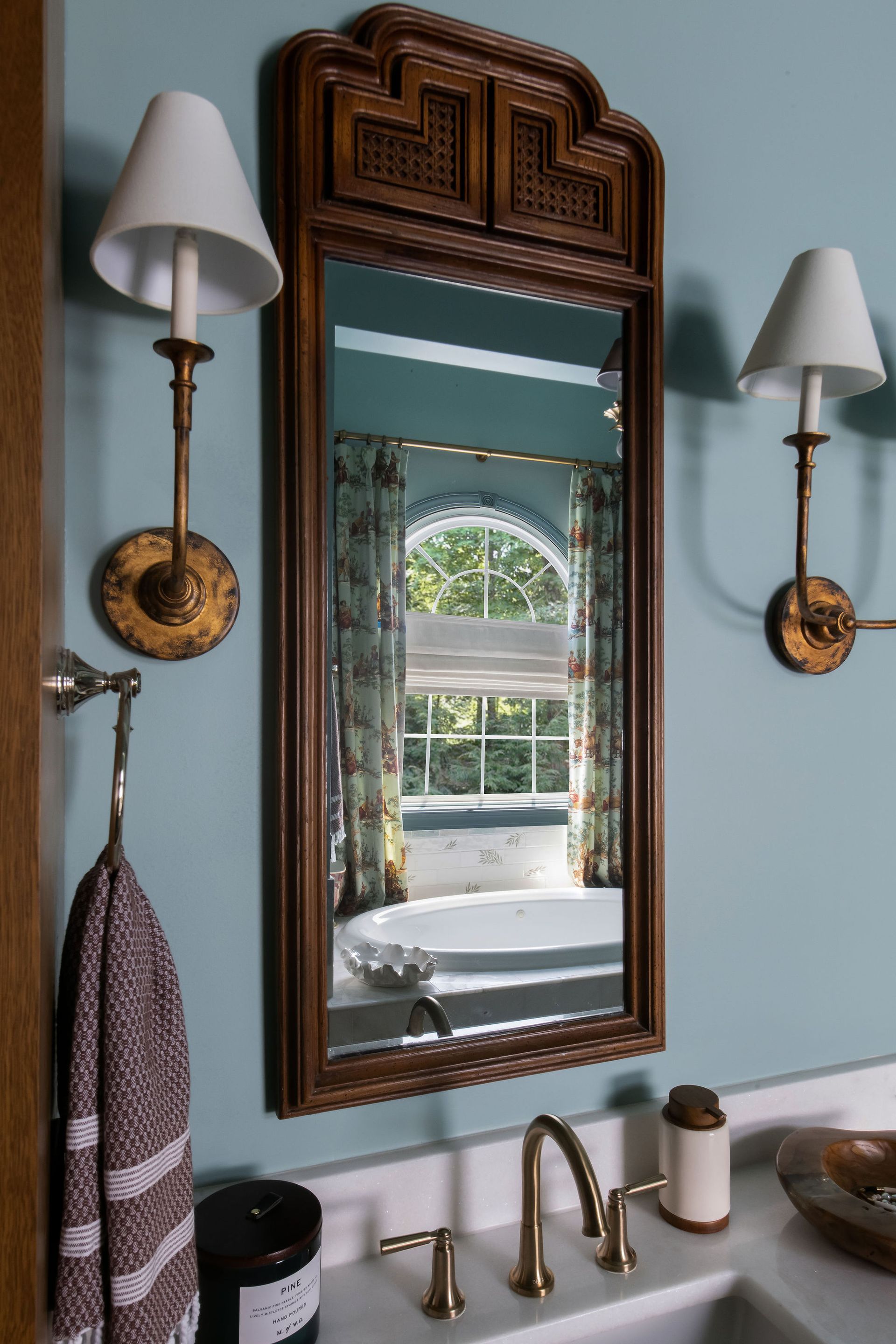 Bathroom with a decorative wooden-framed mirror, sconces, and a view of a window. Blue walls.