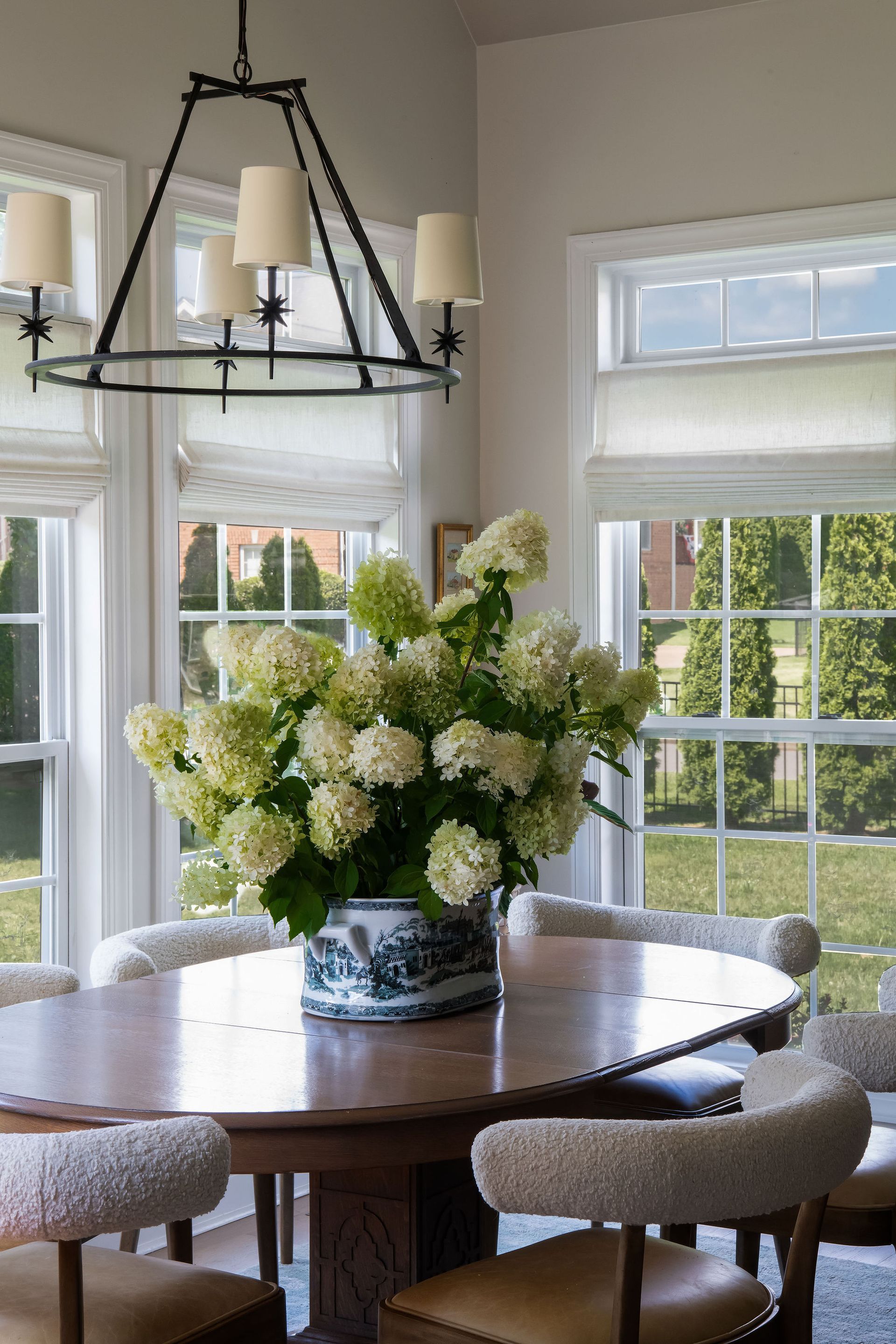 Dining room with round wooden table, chairs, and large floral arrangement, with windows.