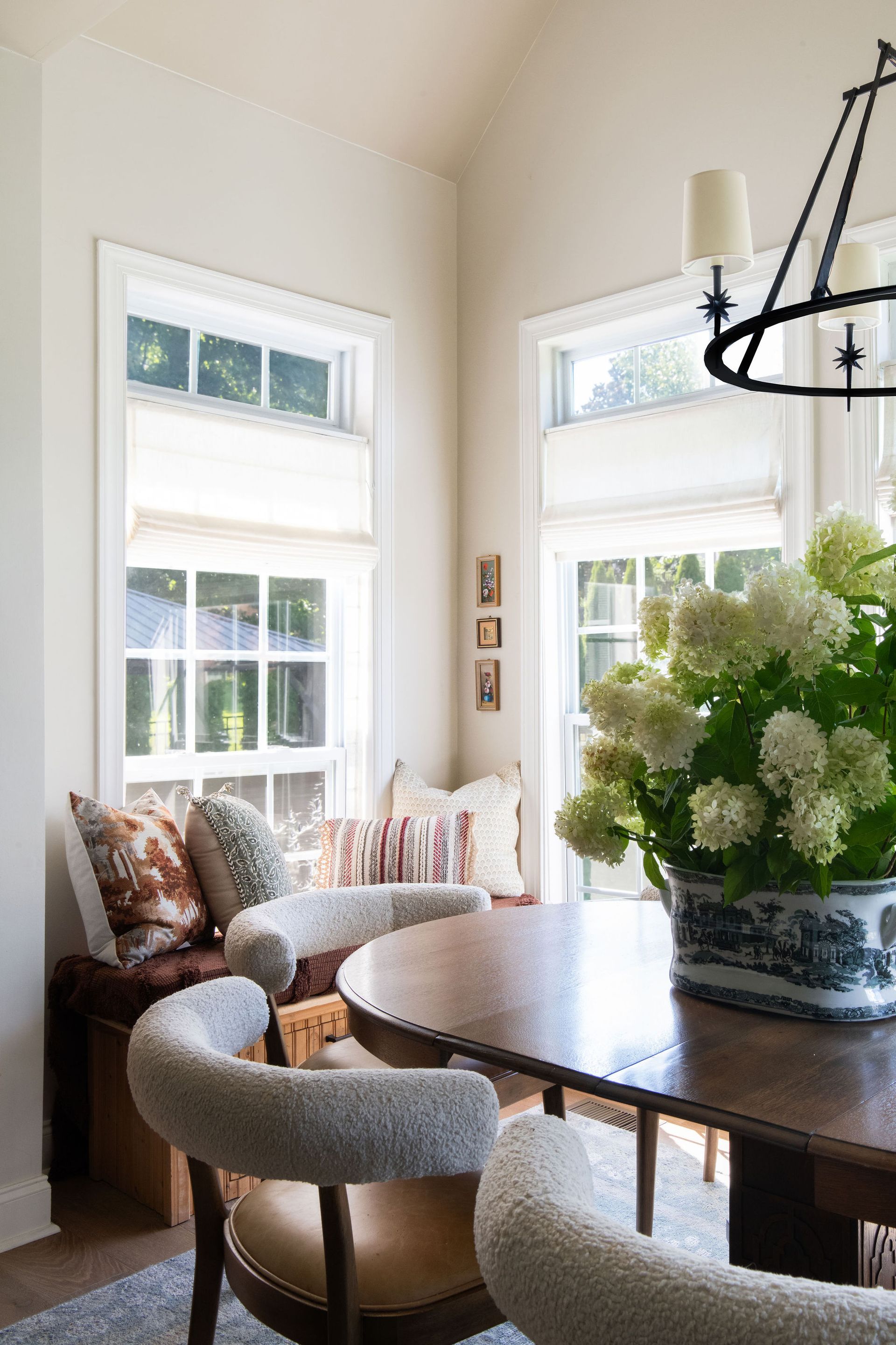 Dining area with round table, chairs, and window seat with pillows; flowers in a blue and white pot.