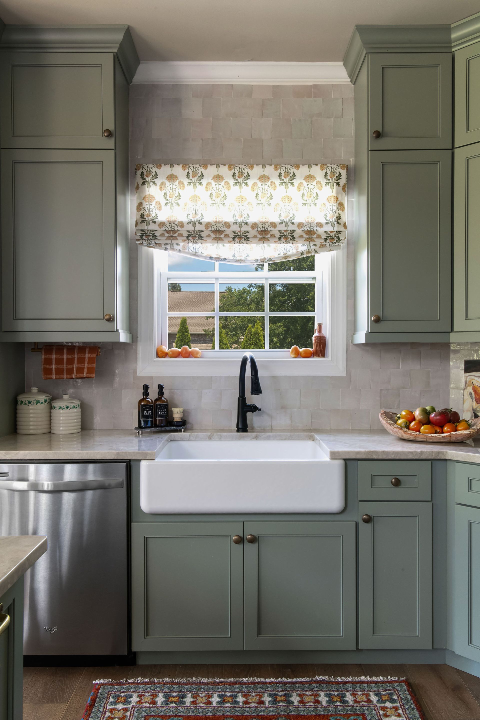 Sage green kitchen with a white farmhouse sink, window with patterned shade, and cabinets.