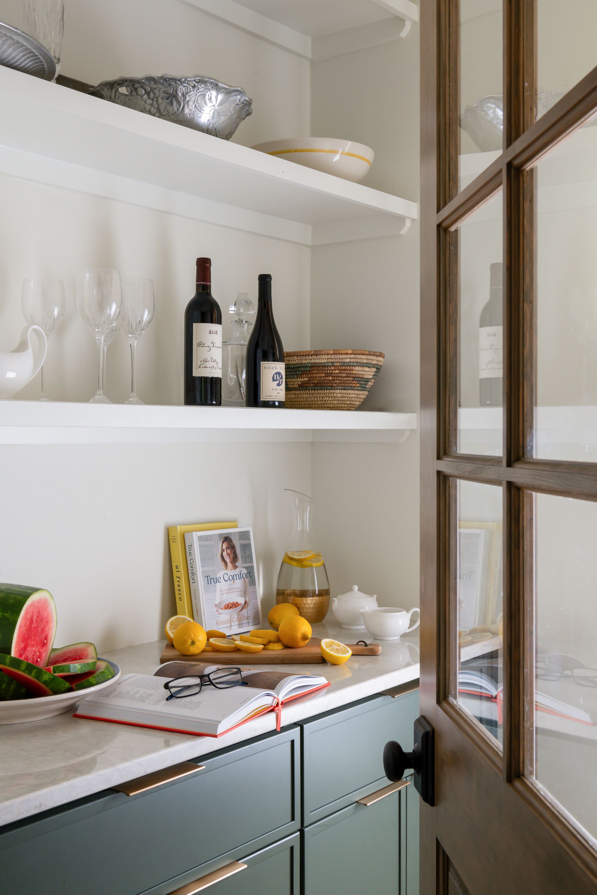 Pantry with open door: wine, glassware, fruit, cookbook, and white shelves against green cabinetry.