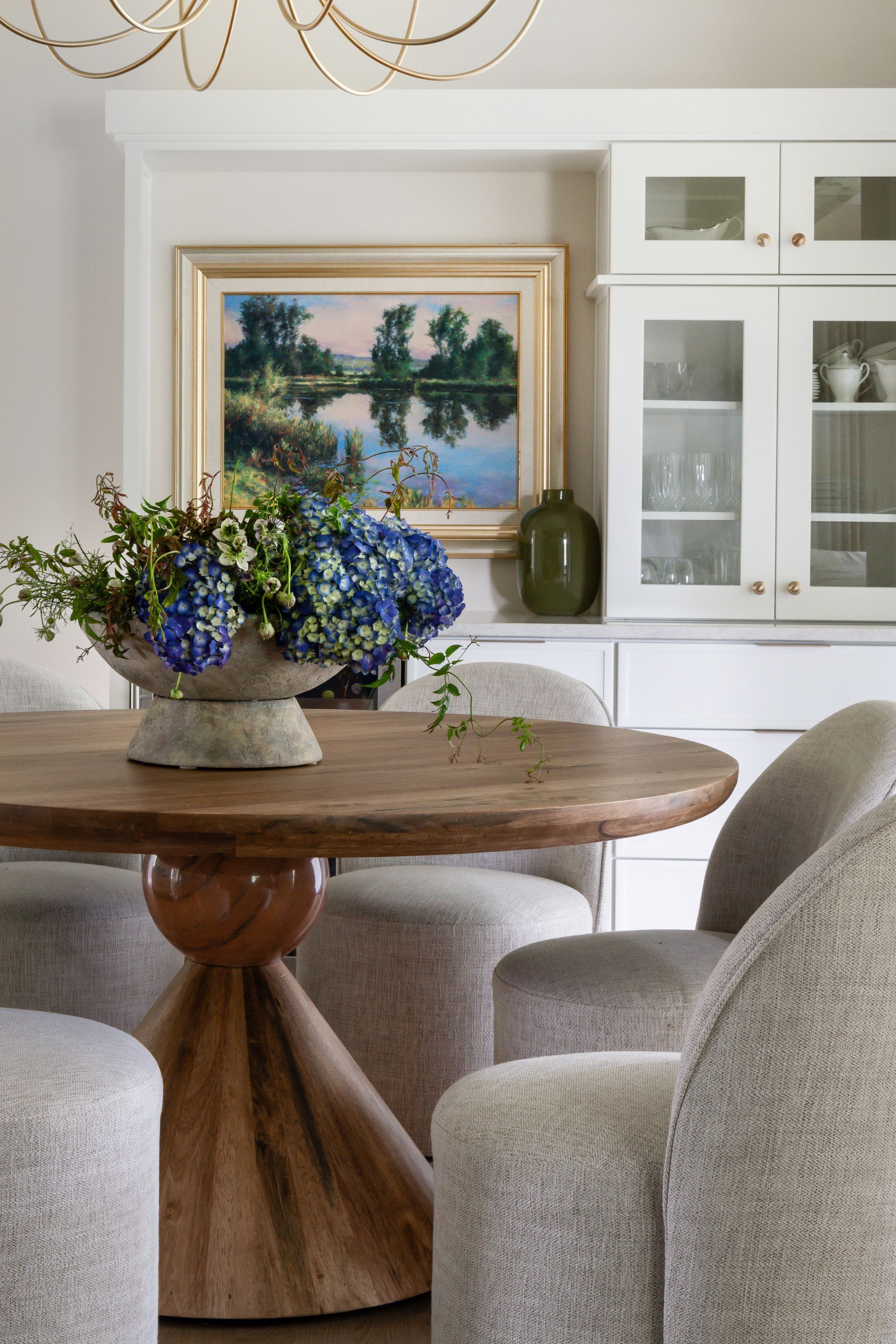 Round wooden dining table with blue floral centerpiece, surrounded by gray upholstered chairs.