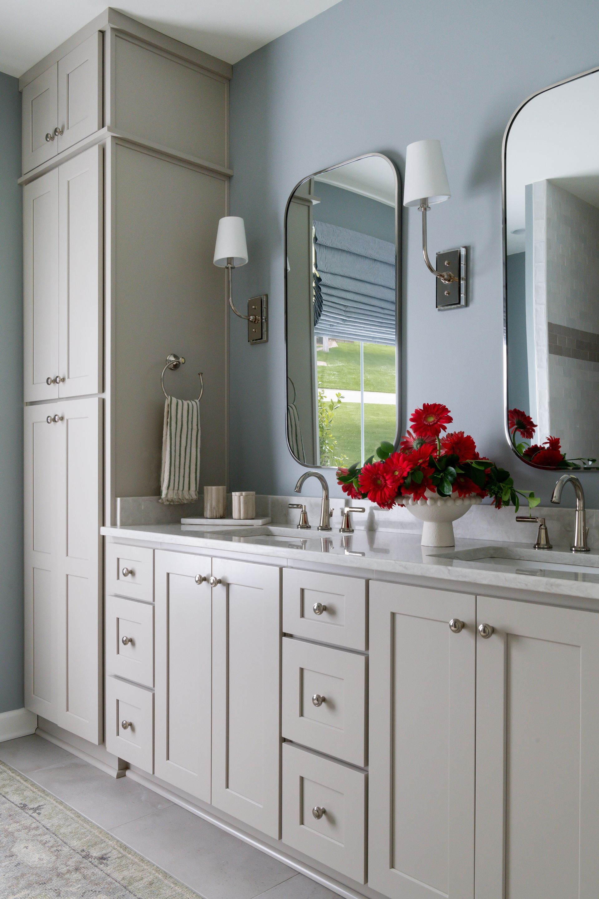 A light blue bathroom with gray cabinetry, two mirrors, and red flowers on the countertop.