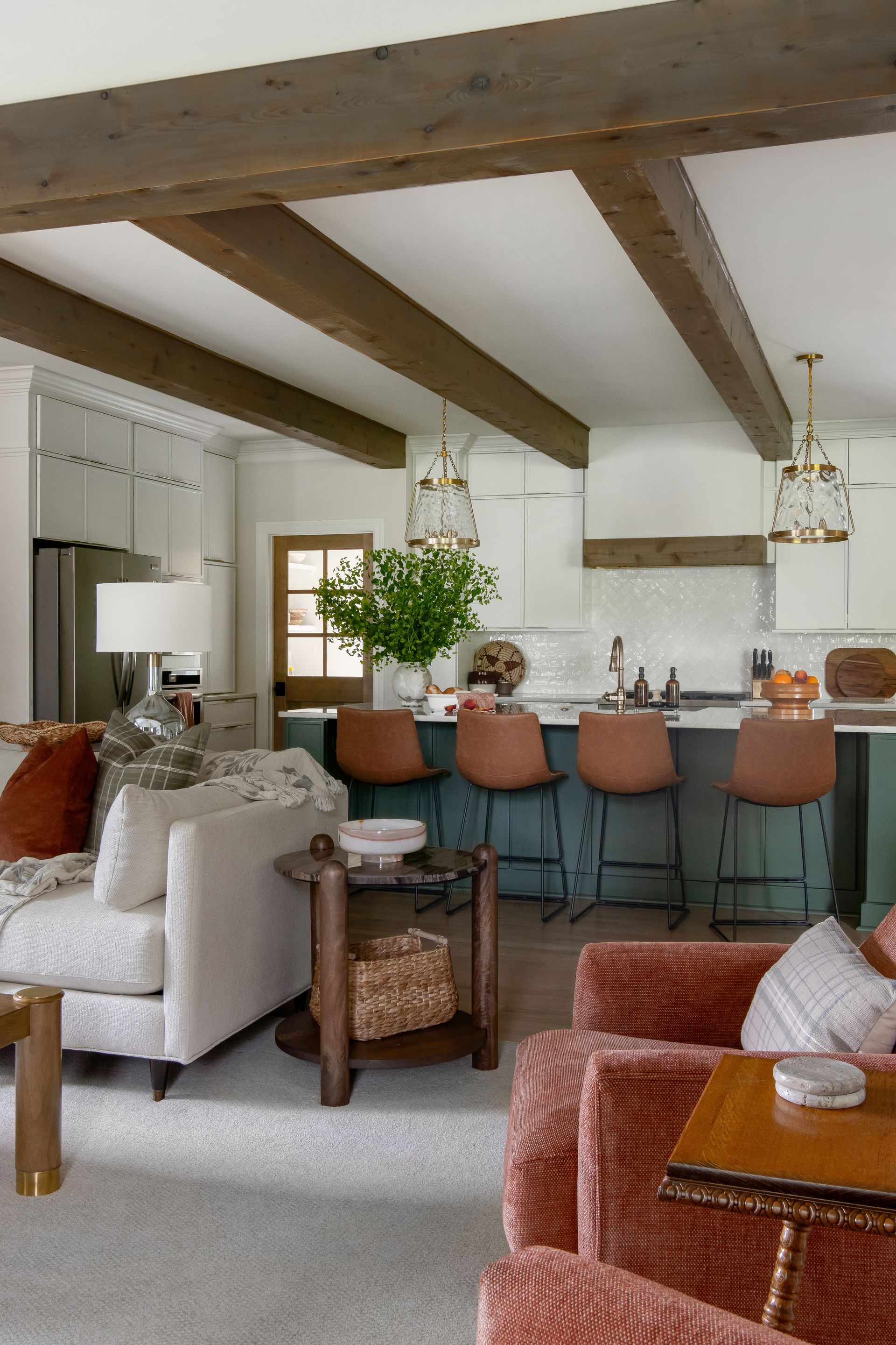 Living room with kitchen. White walls, brown beams, teal island with brown bar stools, tan sofa, and red chairs.