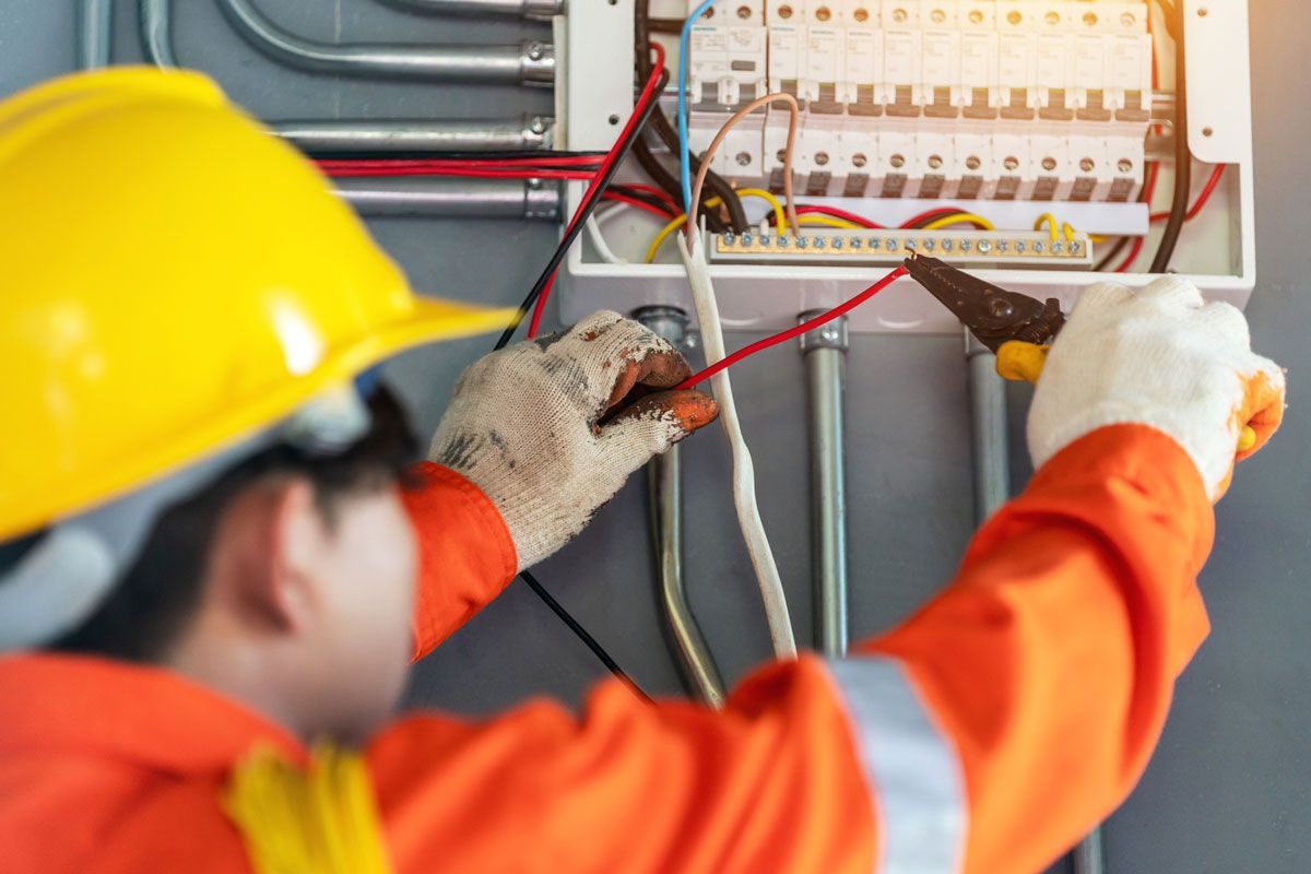 Electrician in orange coveralls and hard hat working on a circuit breaker box, using wire strippers.